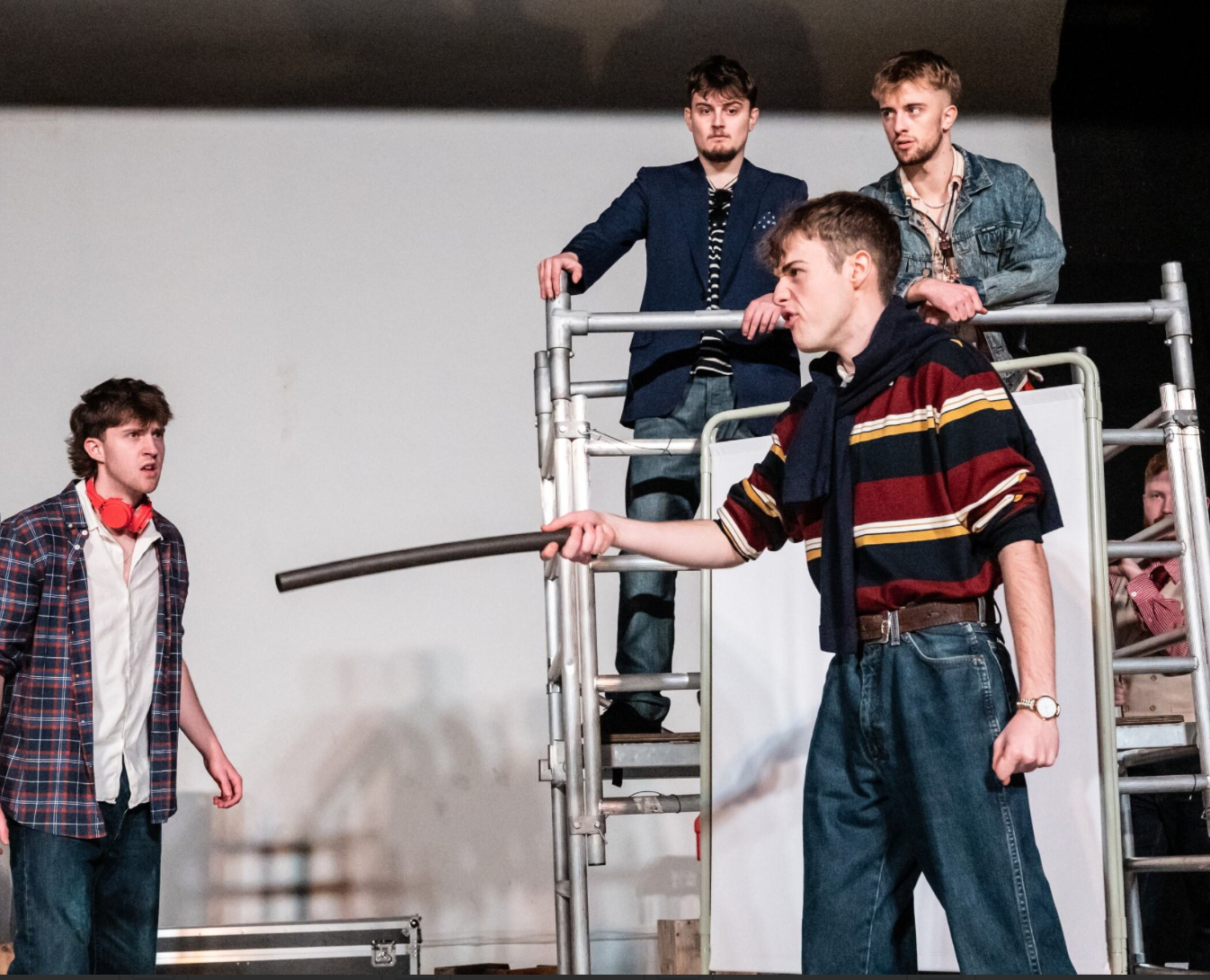 Four BA (Hons) Acting students in modern dress performing in Romeo and Juliet. One of the actors is pointing a piece of pipe at another actor off camera. Two other actors are standing on a small scaffolding behind them.