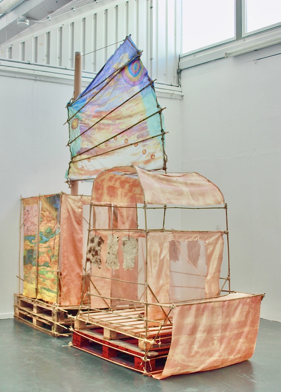 Large installation resembling a Cantonese junk boat, made from bamboo canes, wooden pallets, wire, and twine, with surreal landscape fabric paintings in the shape of a sail and cabin walls, and handmade paper hanging from the structure.