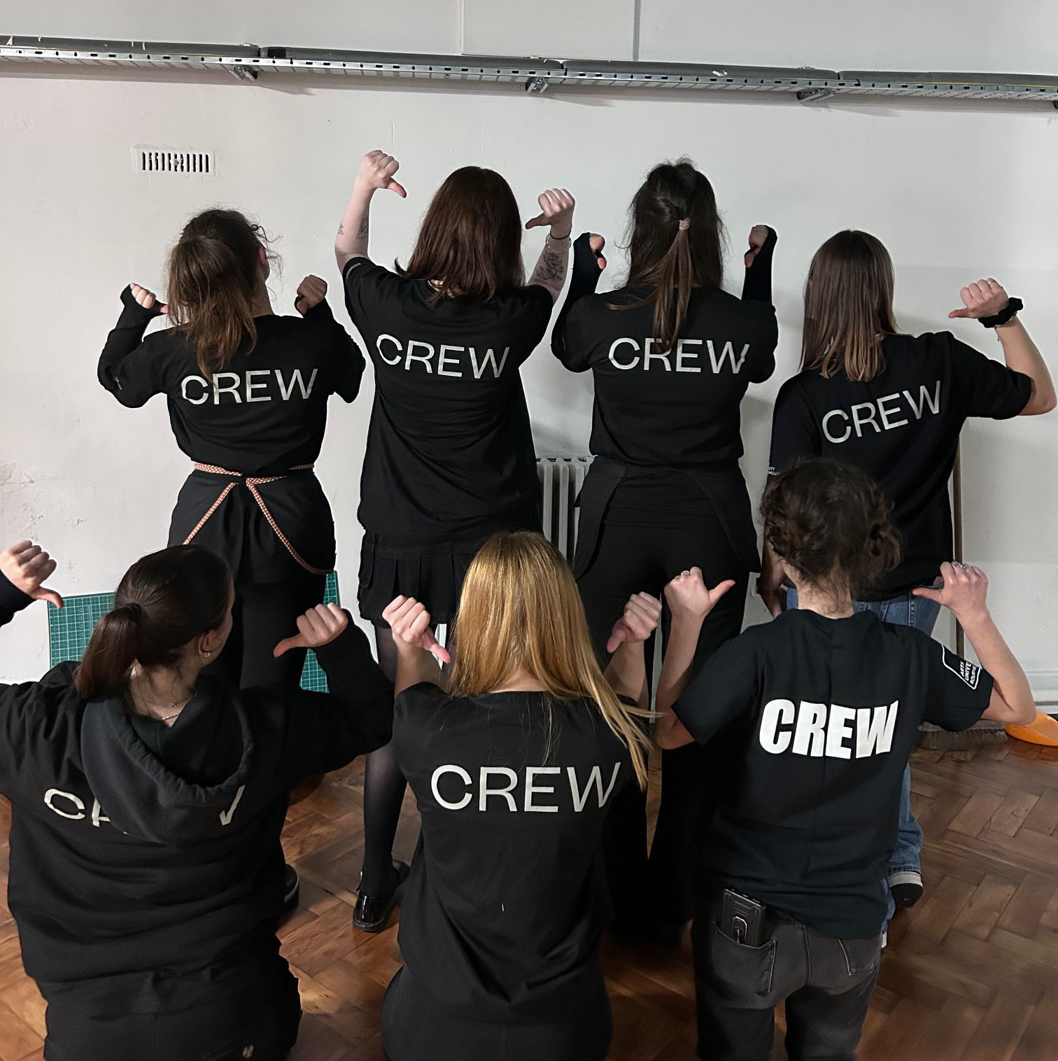 A group of people facing away from the camera pointing at their black shirts which read 'crew'.