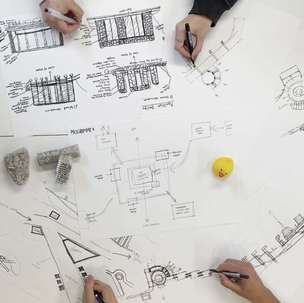 Overhead view of several people working on a series of architectural designs. Pieces of paper are weighted with stones and a rubber duck.
