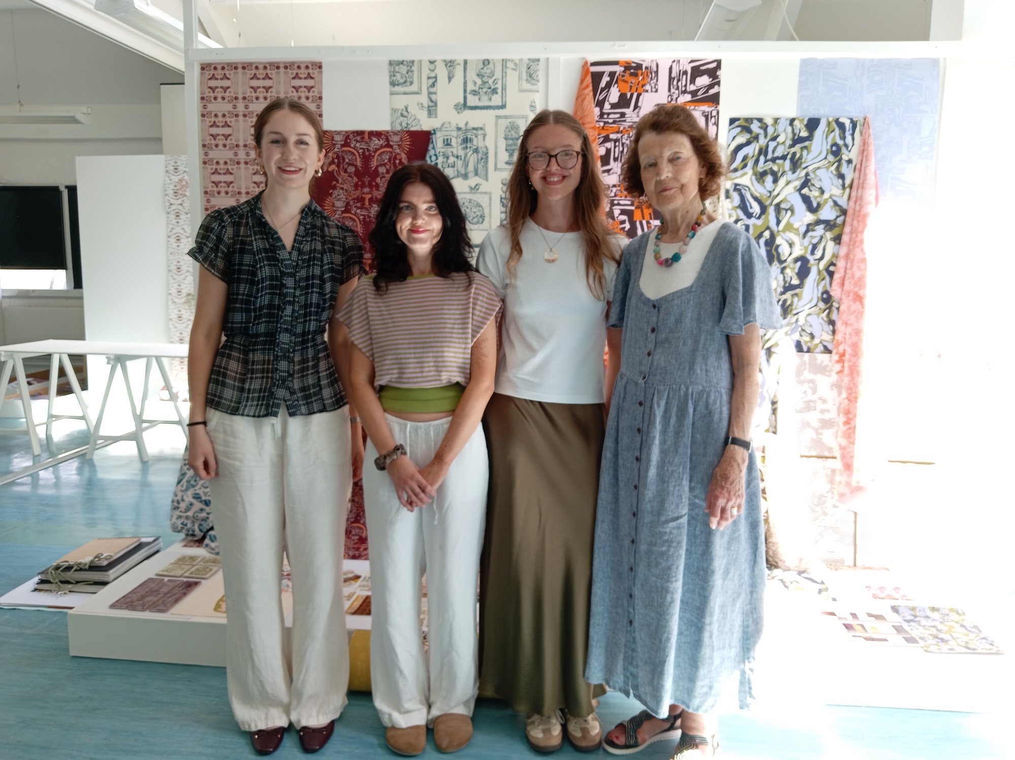 Four people stand together in a brightly lit space with patterned fabric displayed in the background. They are dressed in casual, stylish clothing: a striped top and white trousers, a striped top with a green belt and white trousers, a white top with a skirt, and a blue dress.