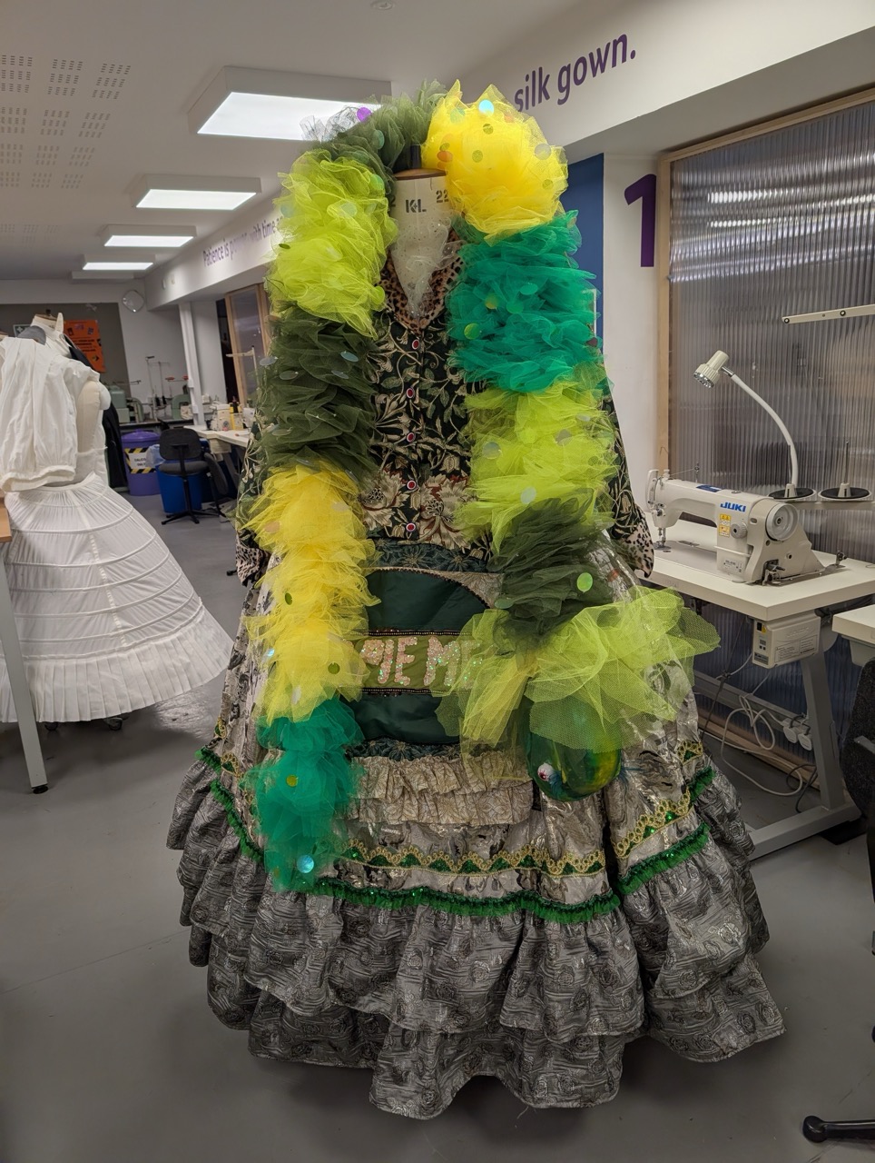 A full-length dress form displaying a vibrant gown with black, white, and multi-coloured elements. The dress features a floral pattern, a frilled skirt, and is adorned with a bright yellow and green tulle overlay. The background includes sewing machines and fabric, indicating a design workspace.