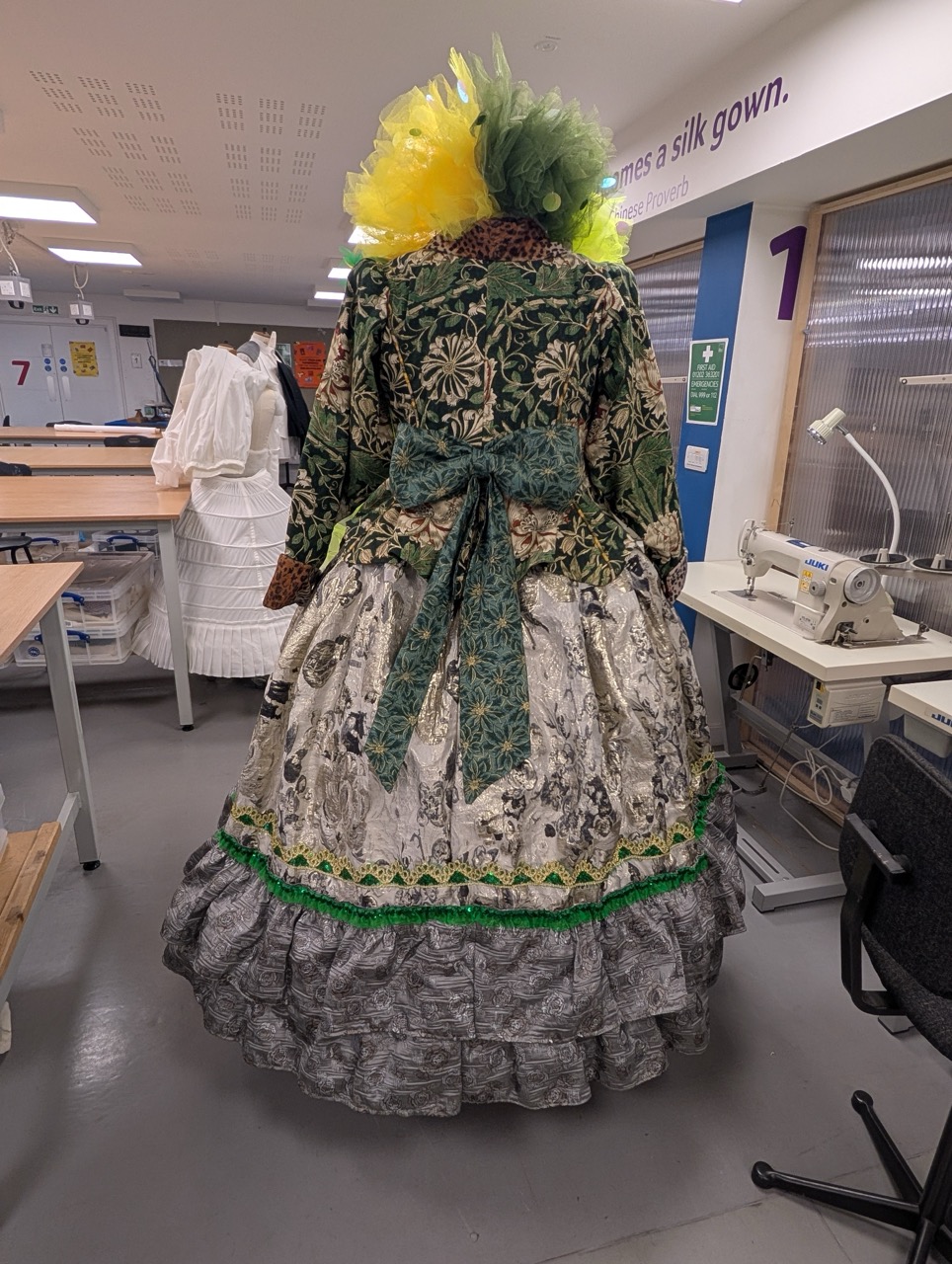 A woman stands with her back to the camera, wearing a large, decorative gown featuring intricate floral patterns in shades of green and grey. The dress has a wide skirt with ruffles and a green bow at the waist, complemented by a vibrant yellow headpiece.
