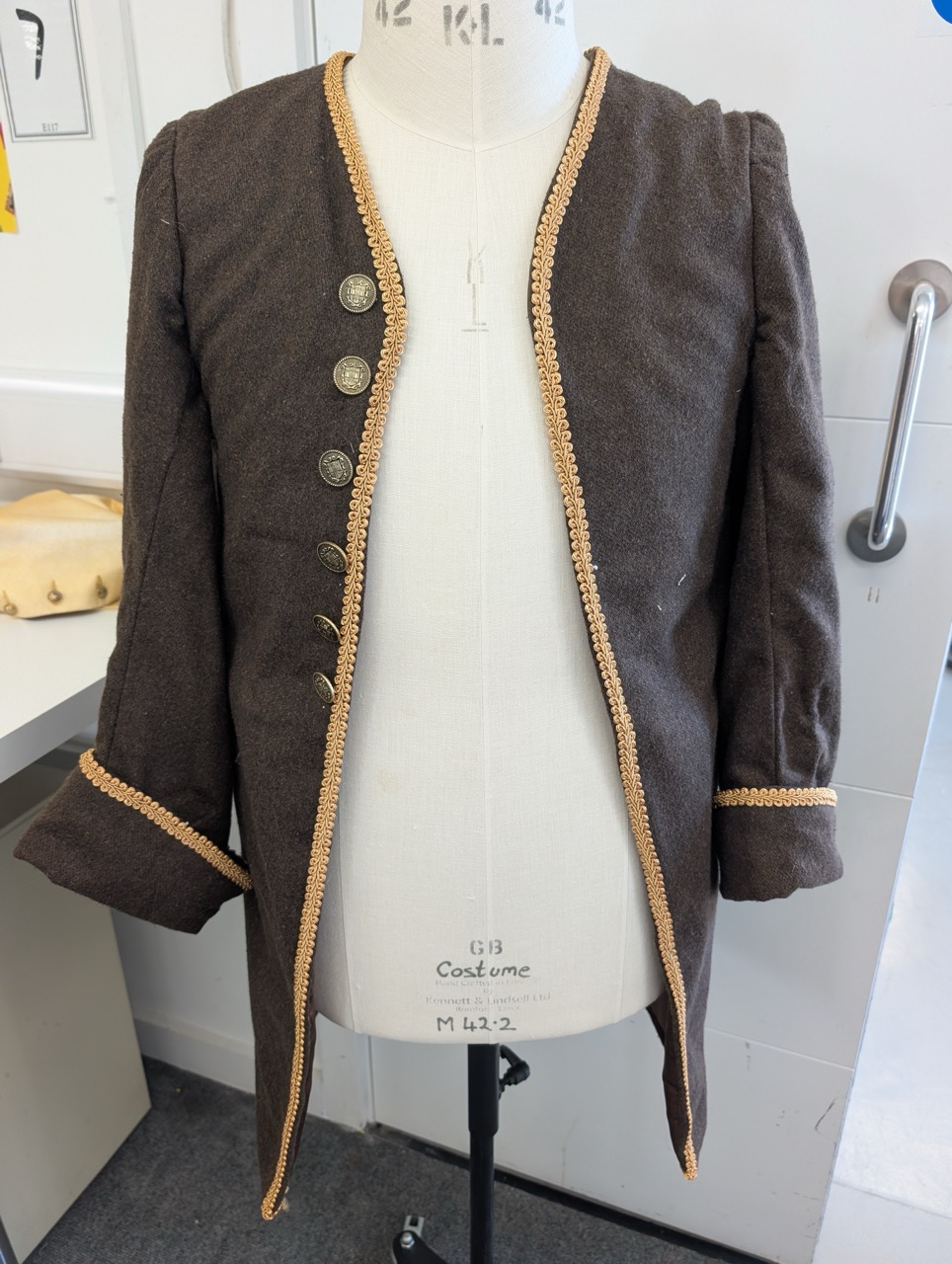 A brown woolen jacket displayed on a dress form. The jacket features a gold trim along the edges and five decorative buttons down the front, with slightly puffed sleeves and angled cuffs.