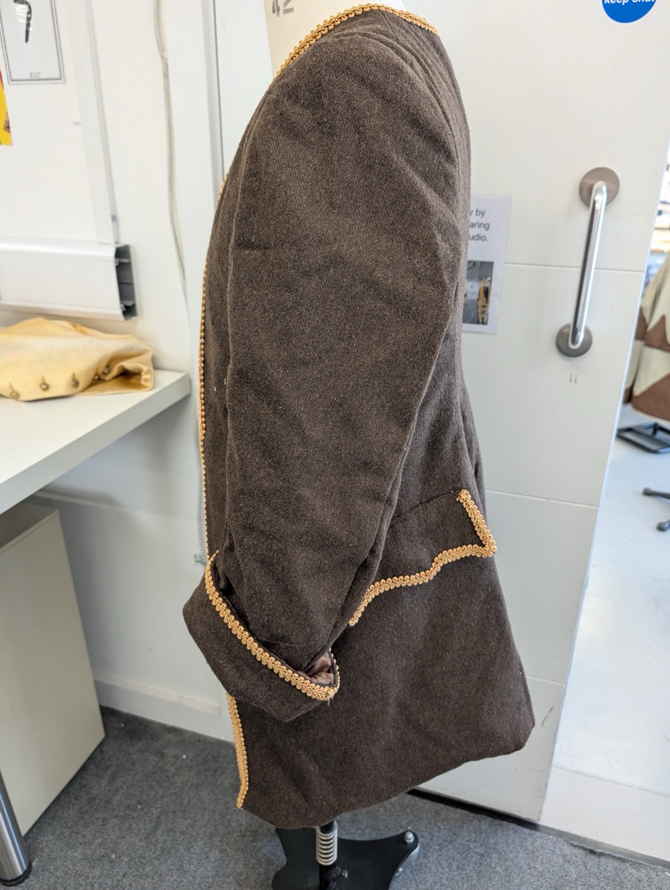 A brown wool coat displayed on a mannequin, showing a side view. The coat features a contrasting trim along the edges and a pocket on the front. In the background, there is a table with a beige fabric and a wall with notice boards.