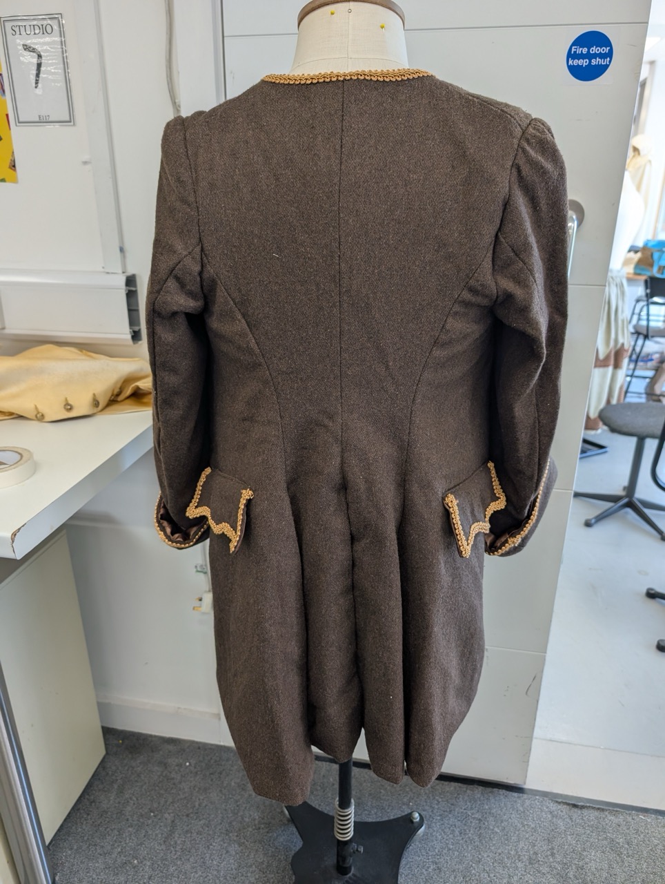 The back view of a brown tailored coat displayed on a dress form. The coat features a fitted silhouette, with decorative trim on the sleeves and pocket flaps, and is situated in a well-lit room with sewing equipment in the background.