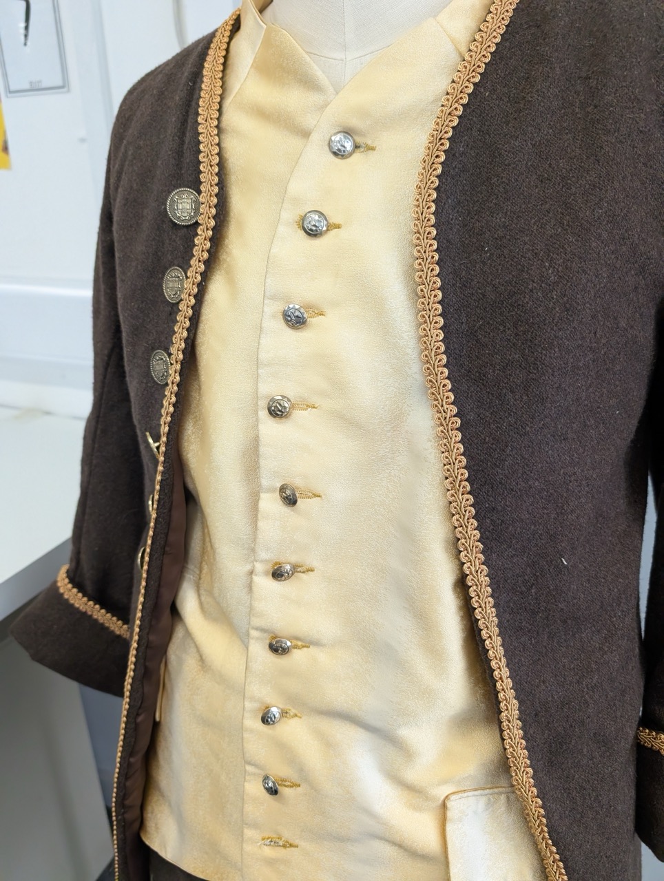 A close-up view of a historical costume displayed on a mannequin. The outfit features a brown jacket with silver buttons and intricate yellow trim, worn over a cream-coloured waistcoat with a row of decorative buttons.