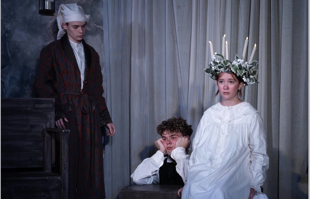 A theatrical scene featuring three actors: a young man in a long robe and hat standing to the left, a young man with curly hair sitting in the middle with his chin resting on his hands, and a young woman in a white dress with a floral crown and candles on her head, seated to the right. The background includes draped fabric and a wooden set.