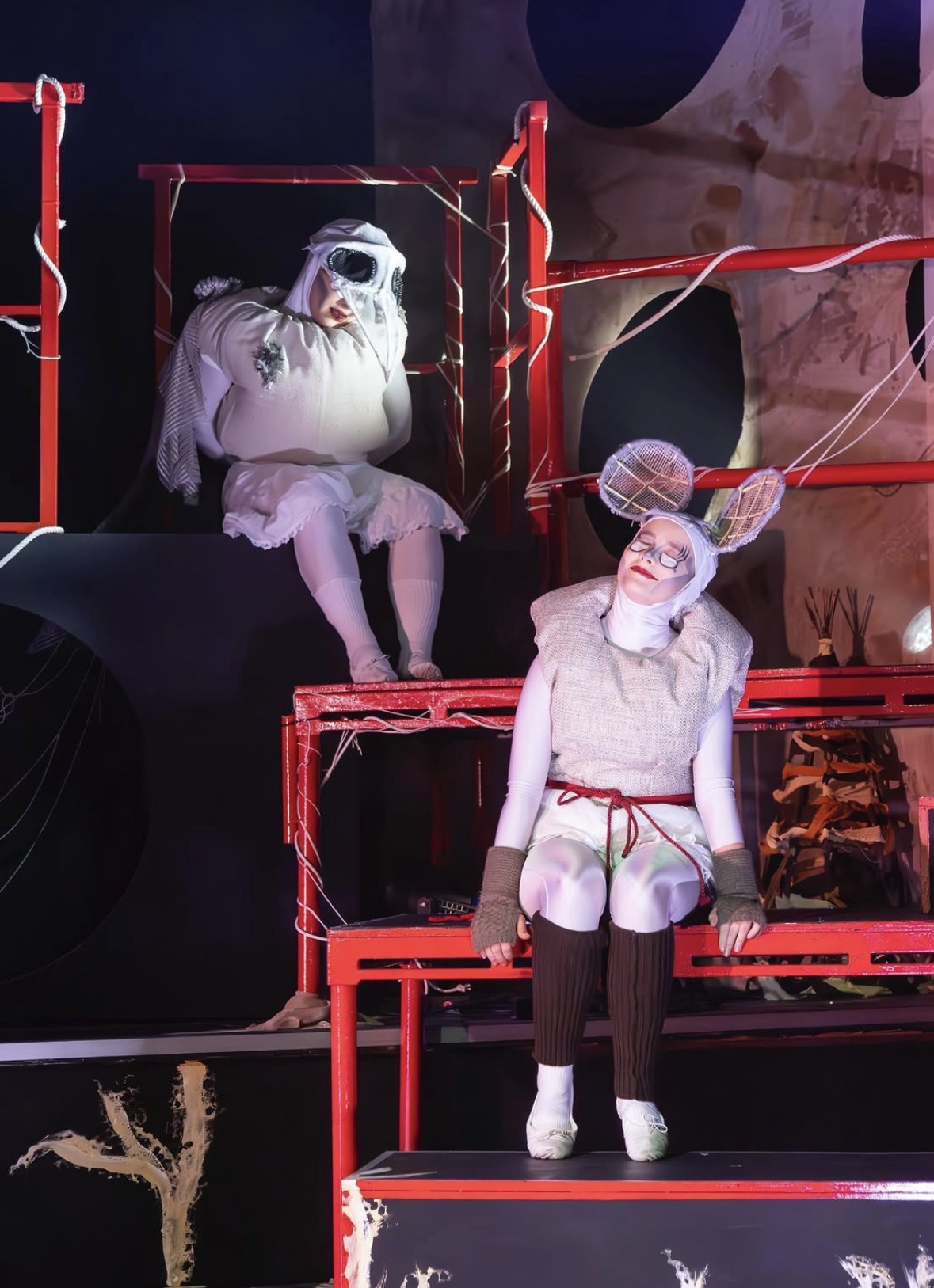 Two BA (Hons) Acting students in elaborate costumes on a stage. One performer, dressed as a mouse, sits on a red structure with shorts and high socks, while another performer, resembling a ghost, reclines in the background. The backdrop features abstract designs.