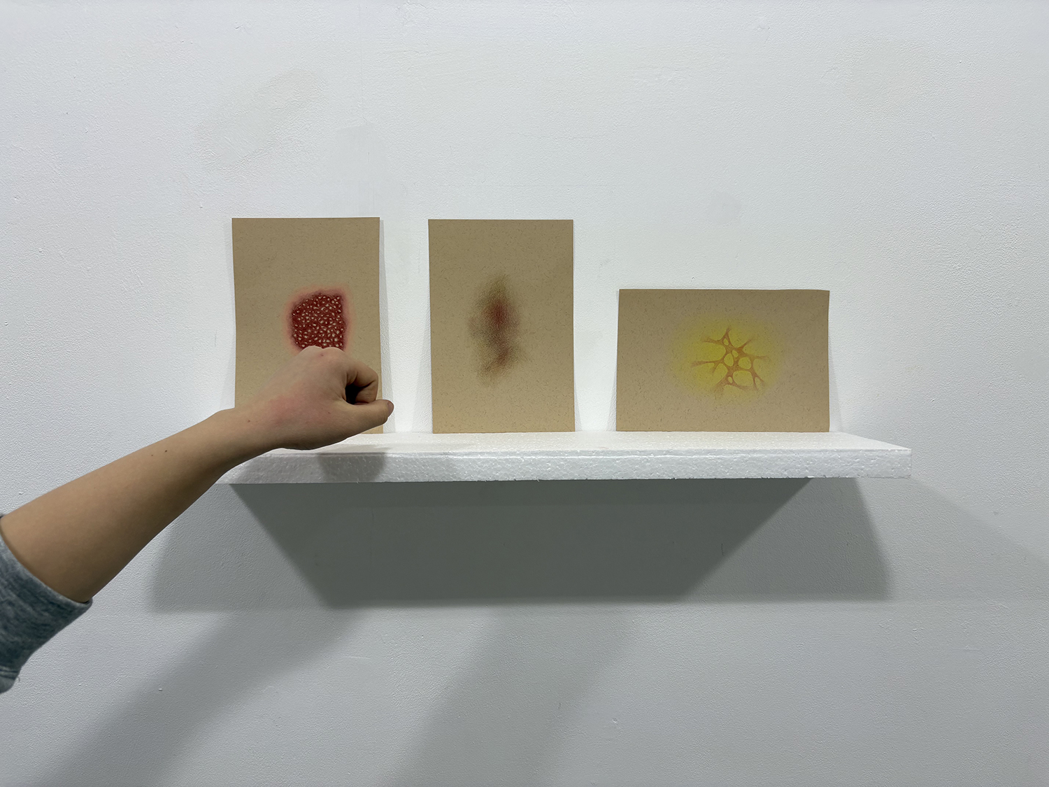 A hand applies red pigment to a small brown canvas on a white wall shelf, beside two other canvases with blurred markings and a yellow star-like drawing.