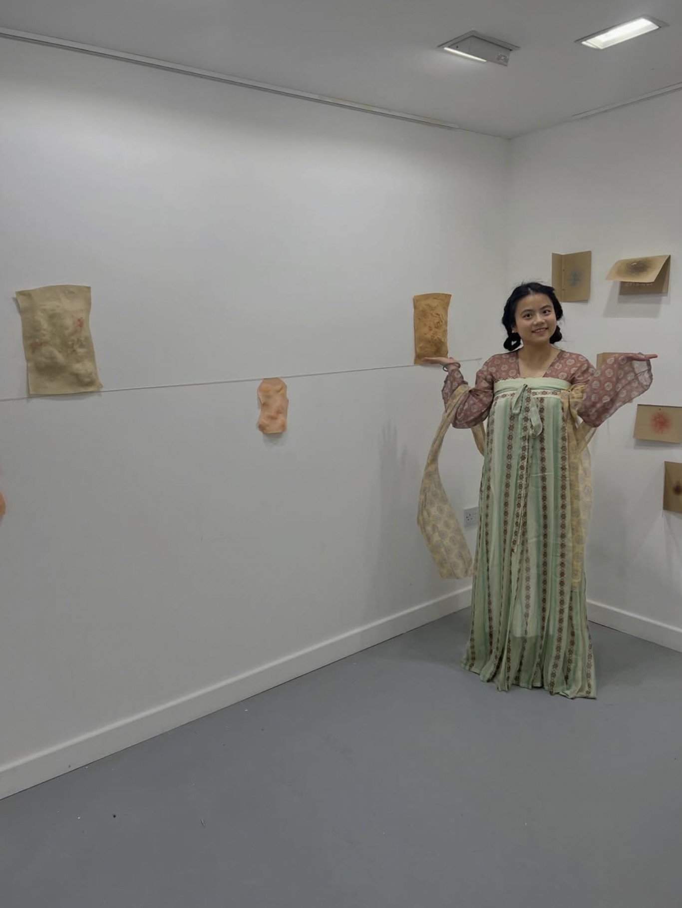 A woman in a long patterned dress stands in a gallery beside wall-mounted textile pieces and small framed works.
