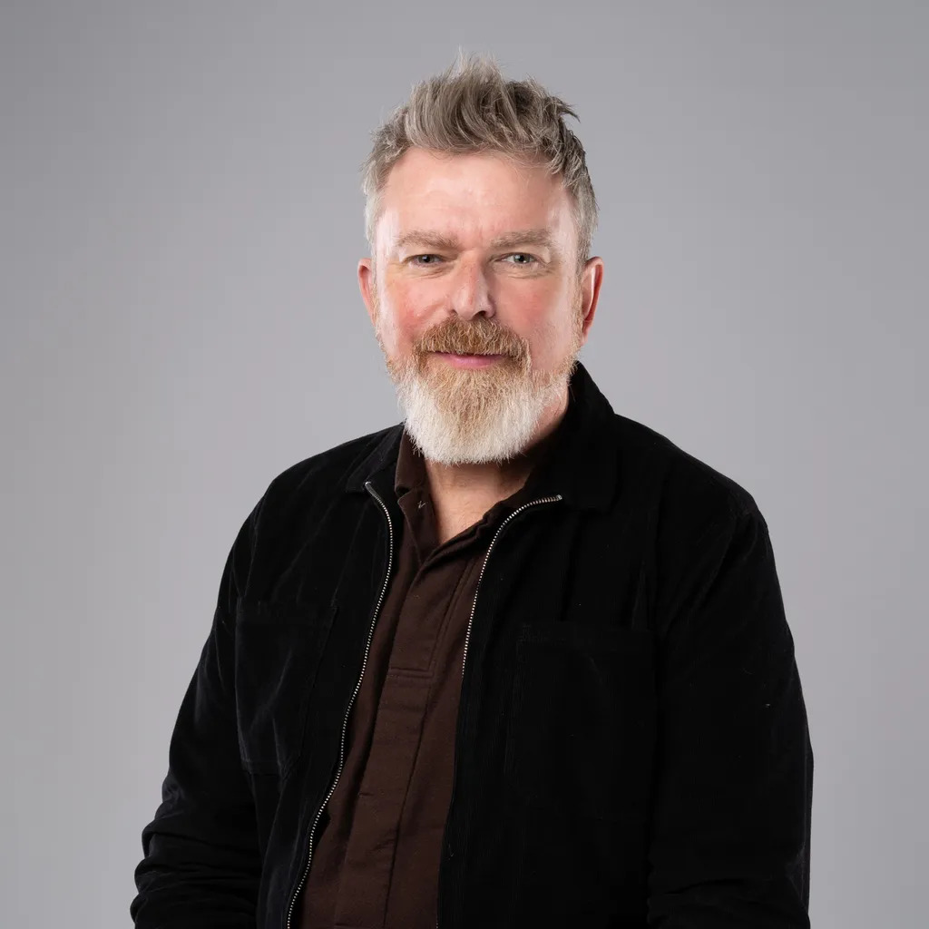 Middle-aged man with blonde hair and grey beard wearing black jacket over brown shirt against grey background.