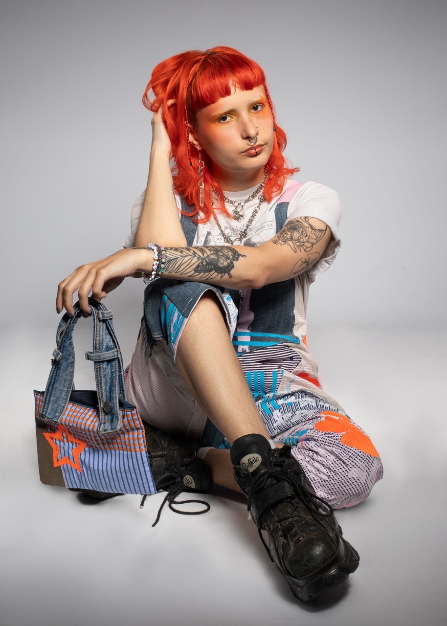 Model with red hair and patchwork-style clothing, a bag made from jean straps and stripy textiles, orange and blue, and large black boots, sat posing, editorial style shot.