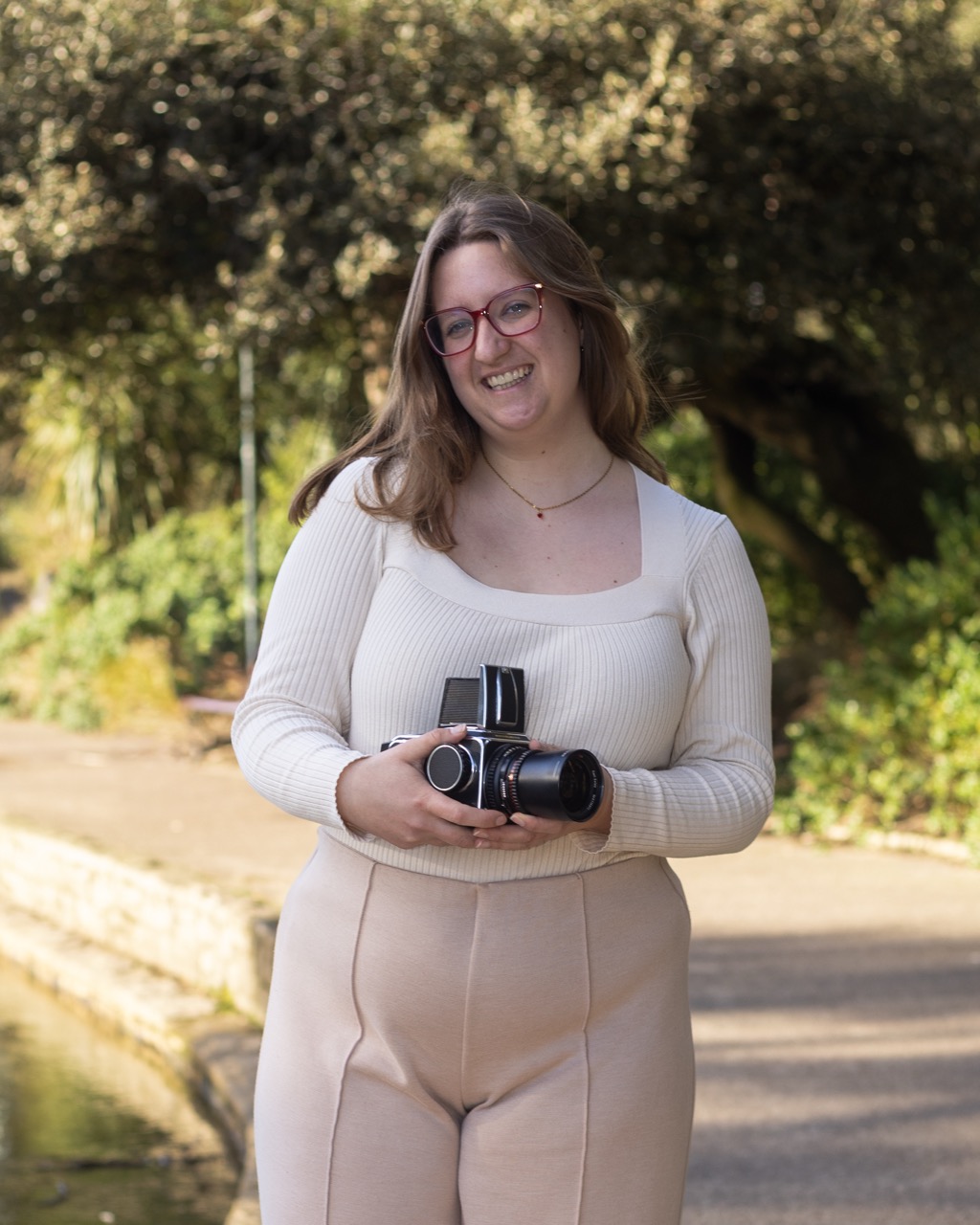 Ruth Virgo stood outside holding a camera, smiling with long hair and red glasses, wearing a necklace and white/beige clothing, foliage and sun in the background.