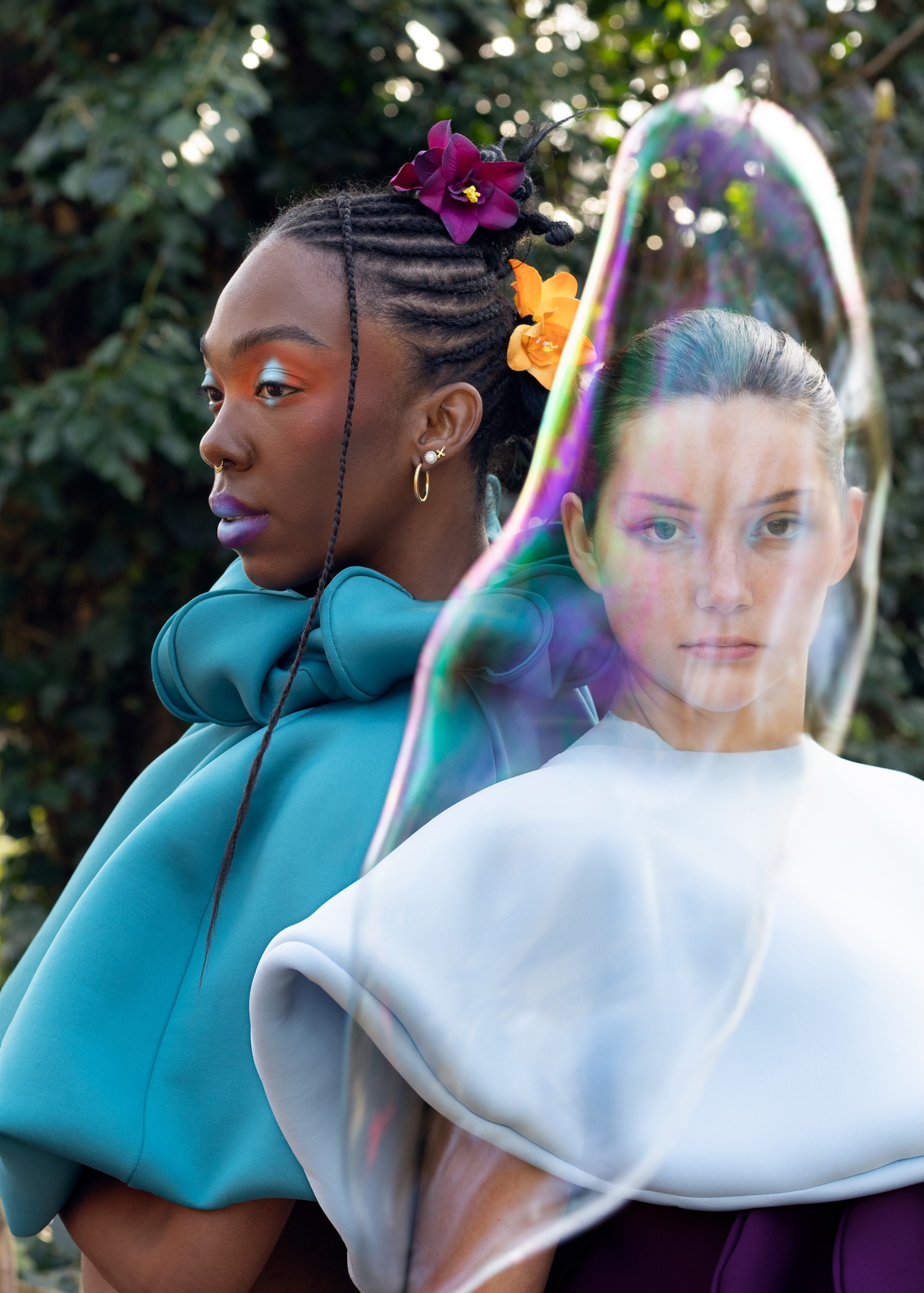 Two fashion models pose for a photo side by side in bold-coloured outfits. A large bubble appears in front of the model on the right.