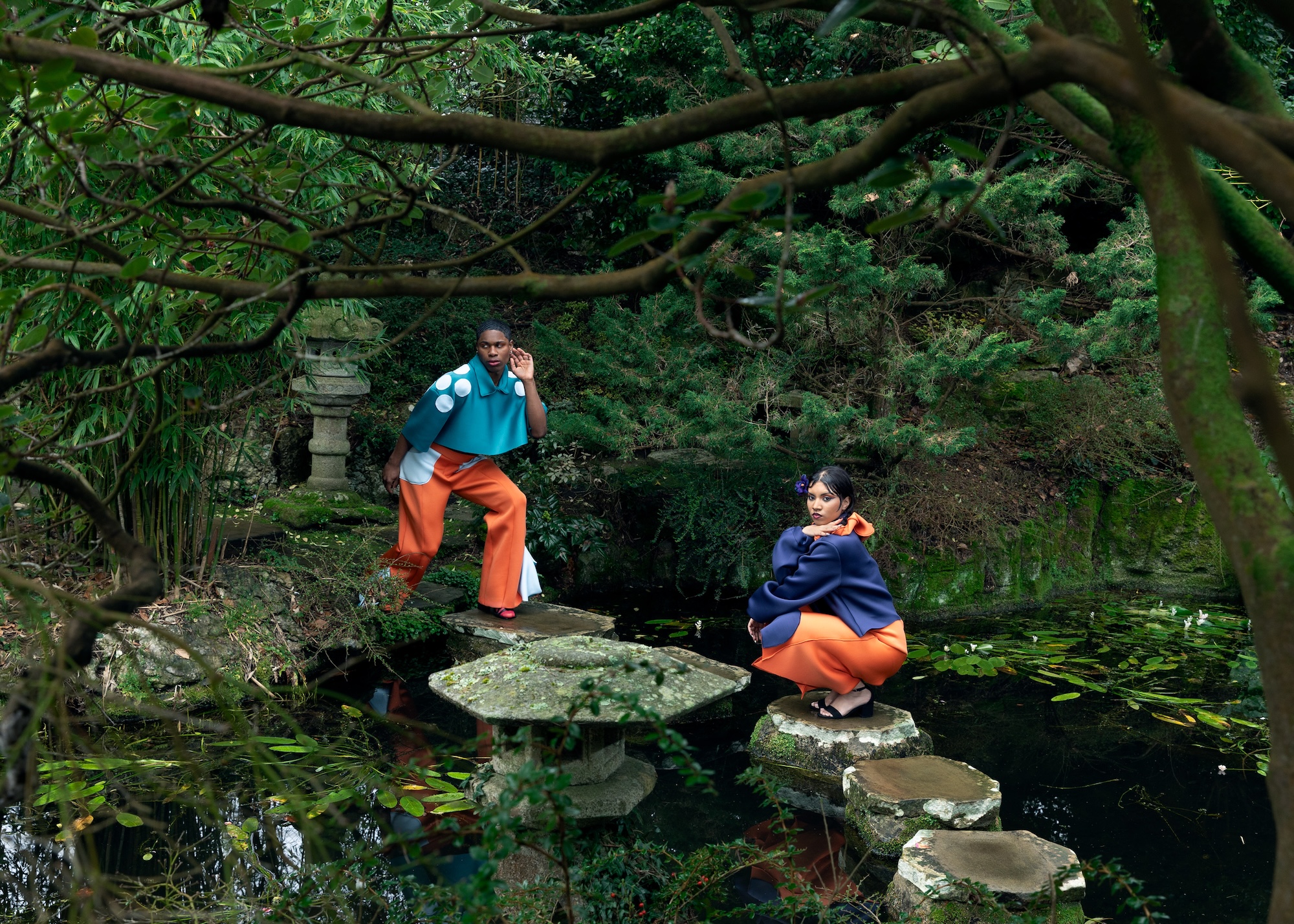 Two fashion models wearing bold blue and orange outfits pose for a photo in a pond in a Japanese-style garden.