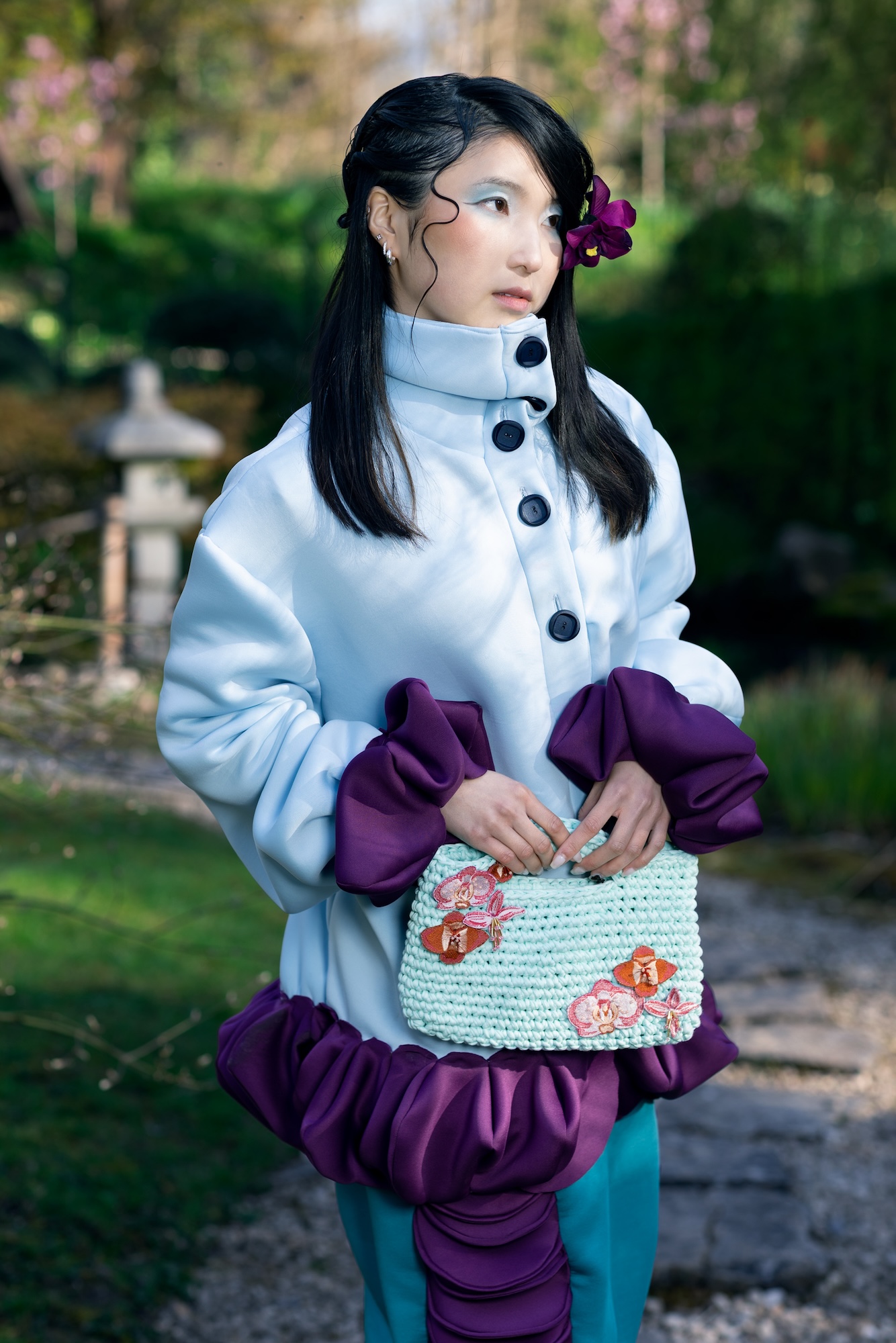 Fashion model wearing a white-grey coat with puffy purple cuffs and accents, holding a floral knitted bag, poses for a photo with a Japanese-style garden in the background.