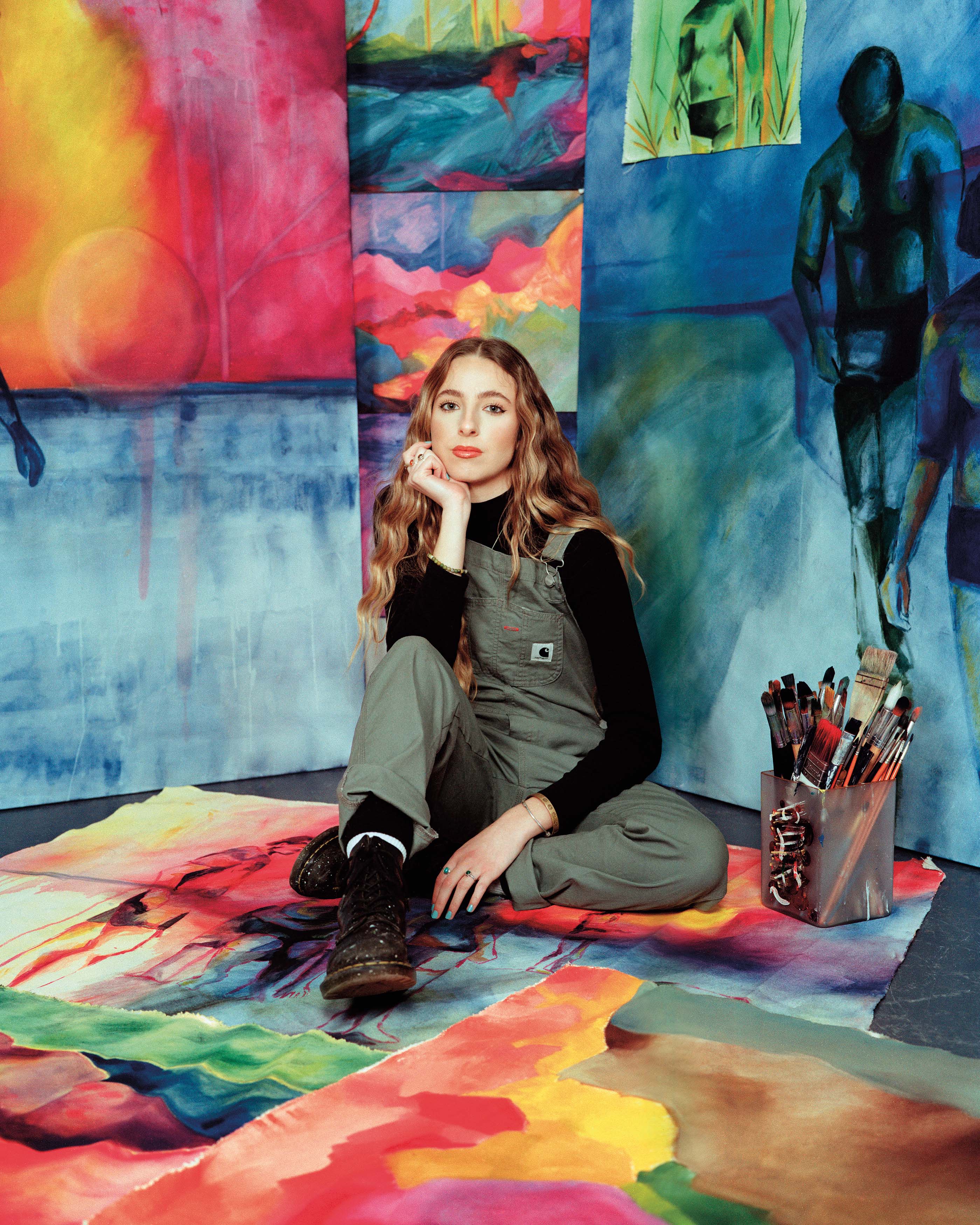 Student wearing grey overalls with long, wavy hair sat amongst multicoloured, abstract artworks.