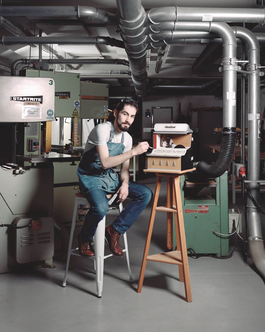 Student with a dark beard and hair wearing denim overalls in an industrial workshop space, painting a wooden and white machine on a wooden chair.