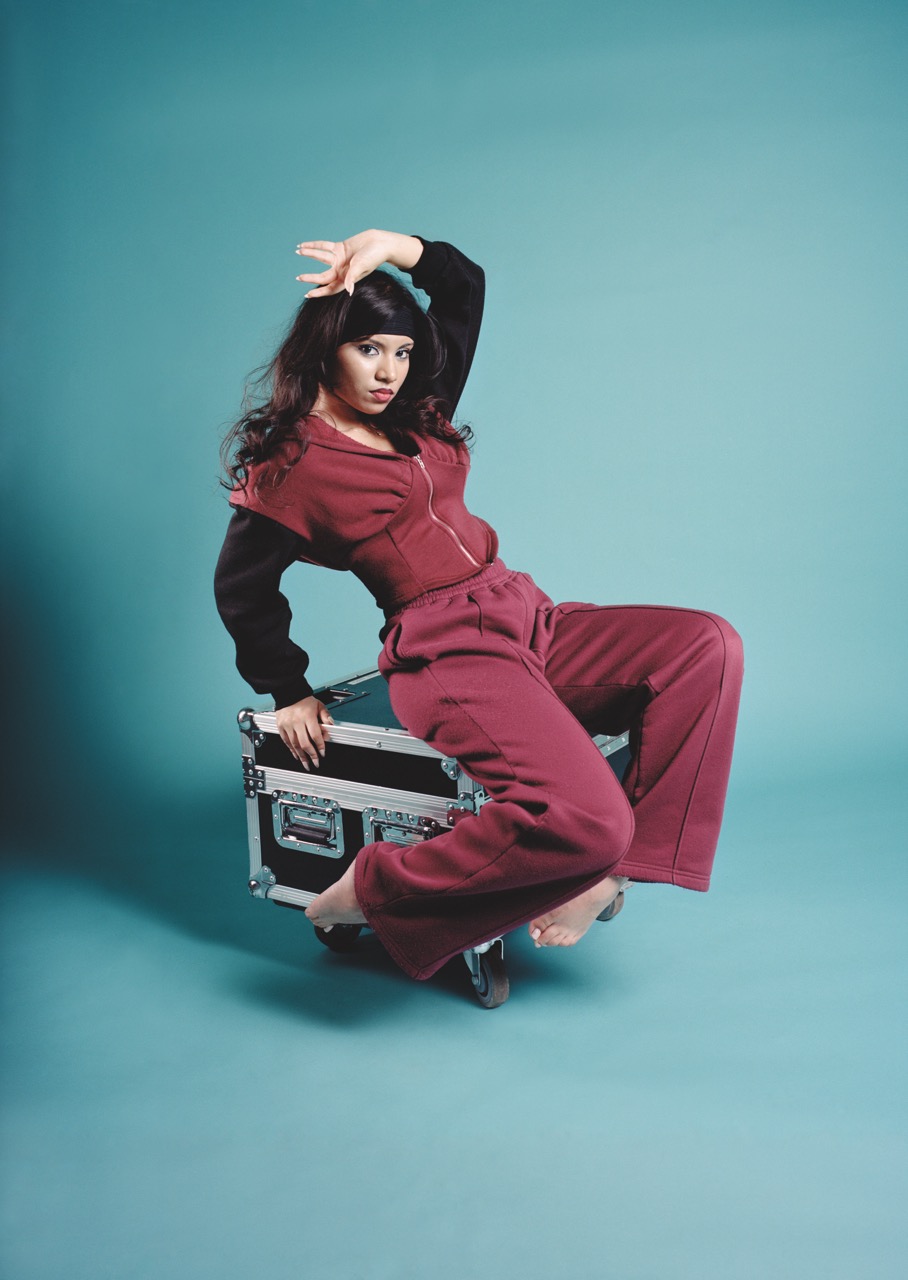 Student in crimson overalls with flowing, dark hair in a dramatic pose on metal stage equipment storage box. Teal studio background.