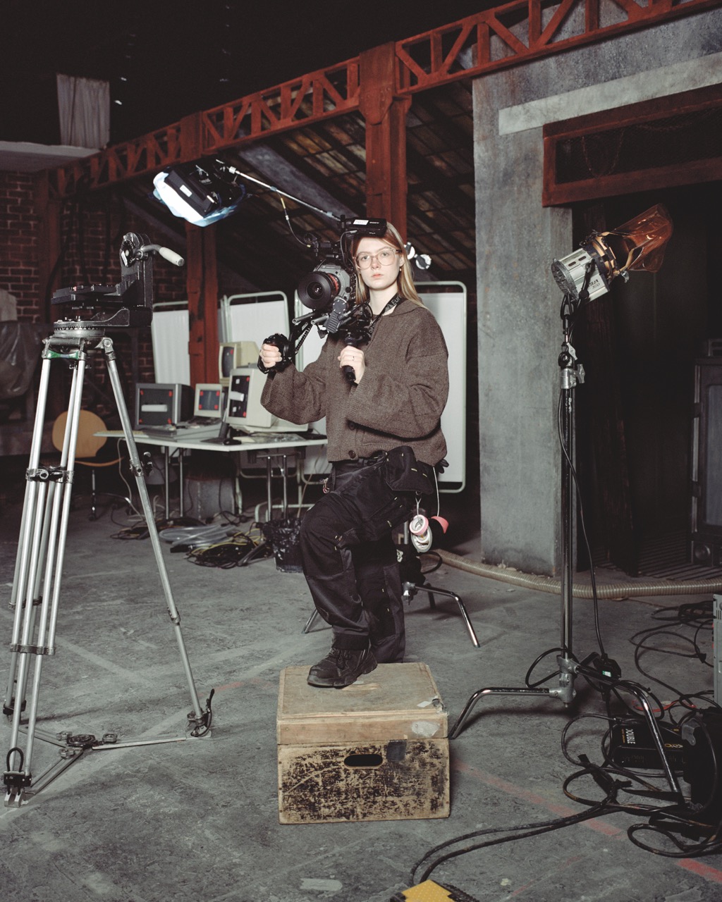 Student with blonde hair and glasses holding heavy camera equipment with a grey jumper and work trousers, posed on a wooden box, surrounded by film equipment on industrial set.