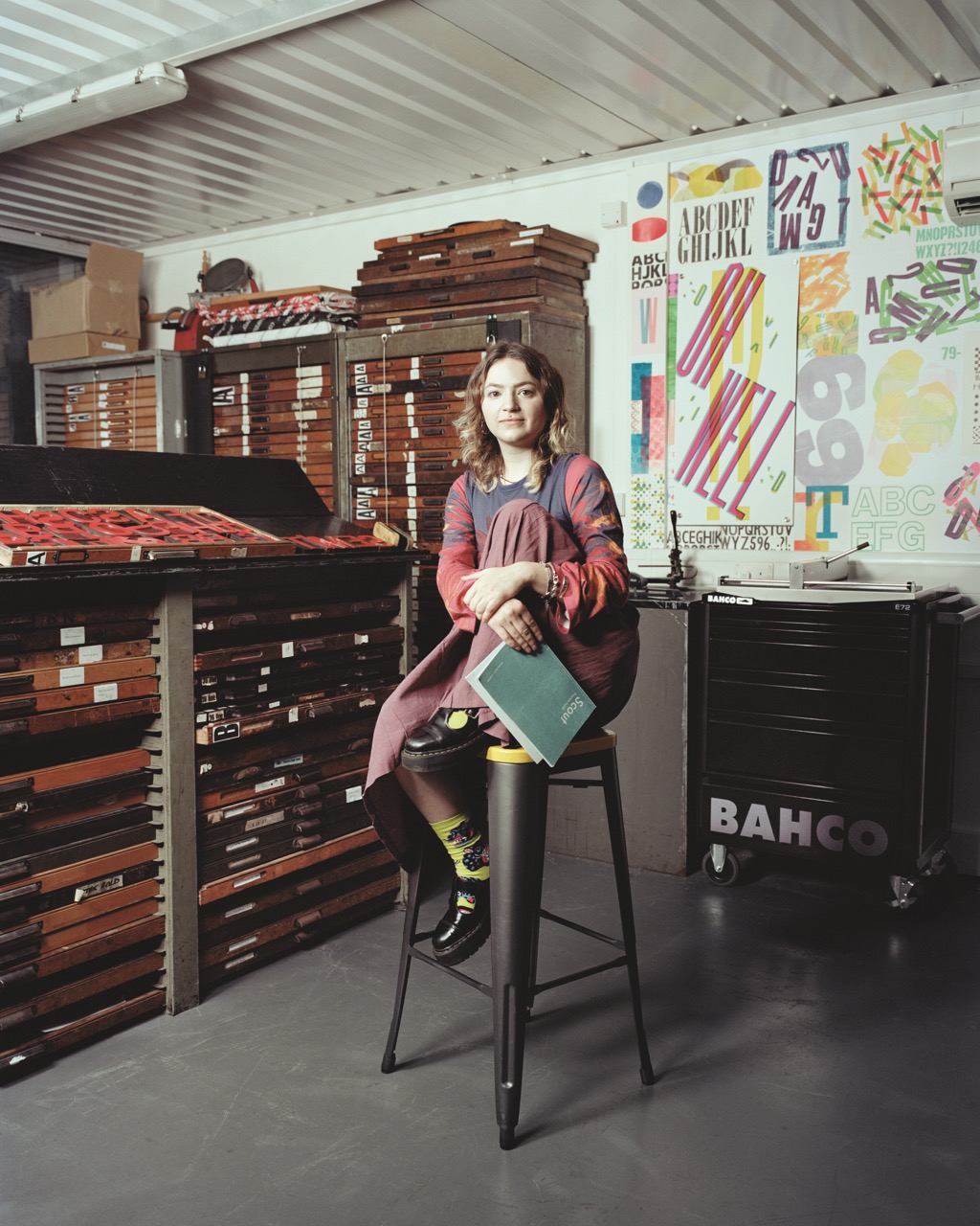 Person sat on a high stool with one leg up, wavy blonde hair and loose blue/red clothing and skirt. They're surrounded by graphic design elements and wooden storage.
