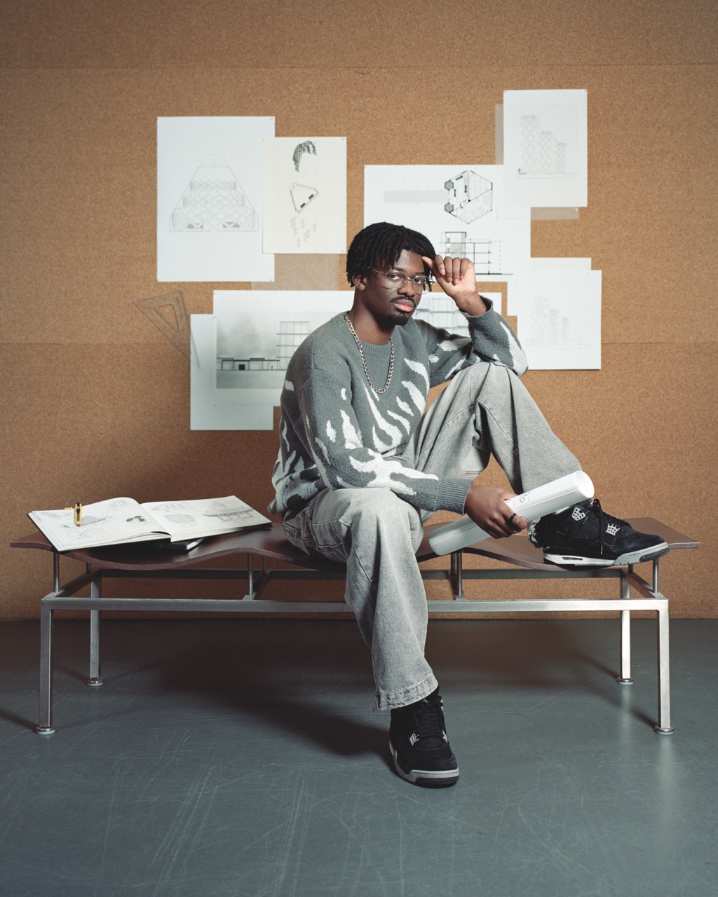 Student sat posing on a long wavy chair with baggy grey sweater and jeans, silver glasses, chain and short locs. White blueprints behind them on a cork wall.