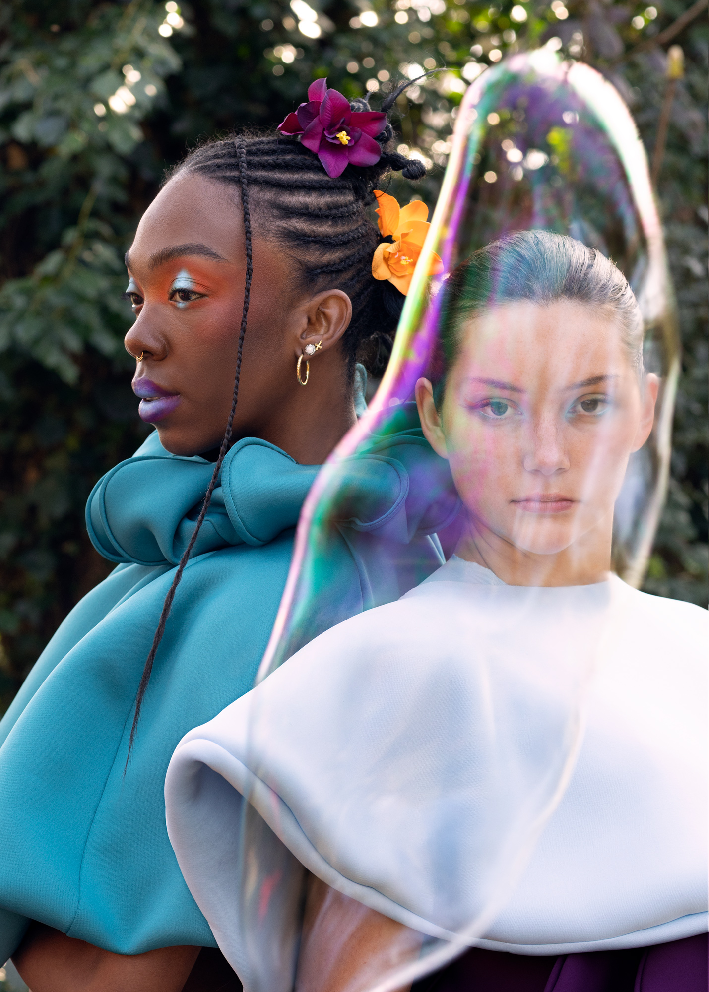 Two models pose in vibrant fashion outfits against a natural background. One model has braided hair adorned with flowers and wears a teal garment, while the other has a sleek look in a white and black outfit, with a soap bubble visually separating them.