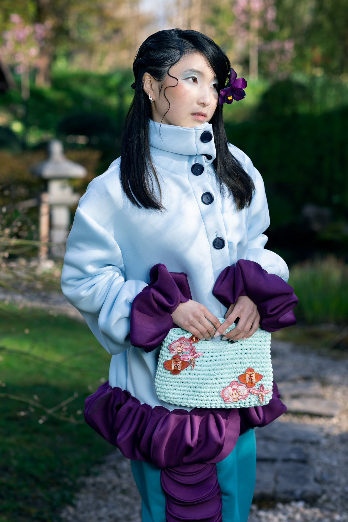 A woman stands outdoors in a garden, wearing a light blue, oversized coat with a purple ruffled hem and large buttons. She holds a mint green handbag decorated with flowers, while her long, dark hair features a purple flower accessory.