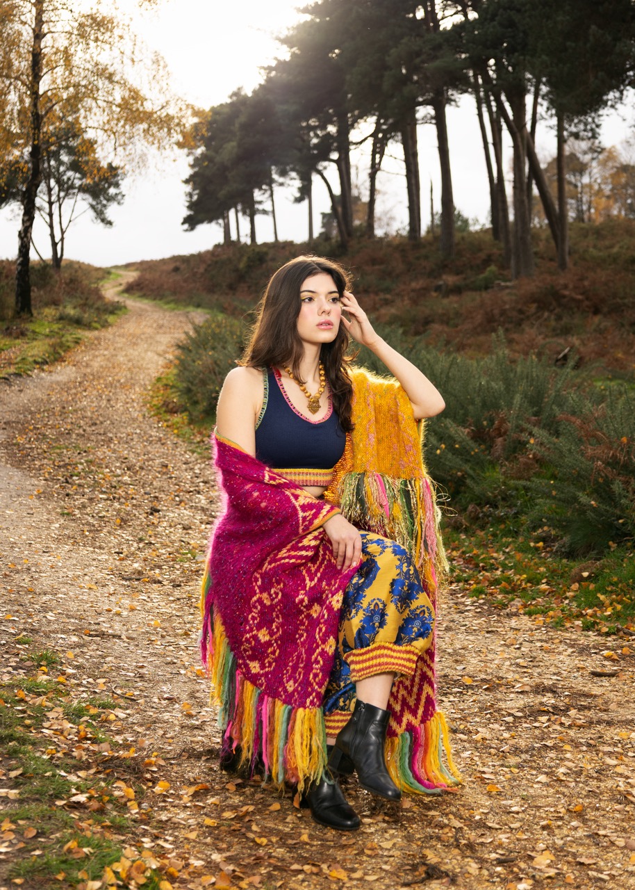 A young woman sits on a path surrounded by autumn foliage. She wears a colourful dress and a vibrant shawl with fringe. The background features trees and a gravel path leading into a wooded area.