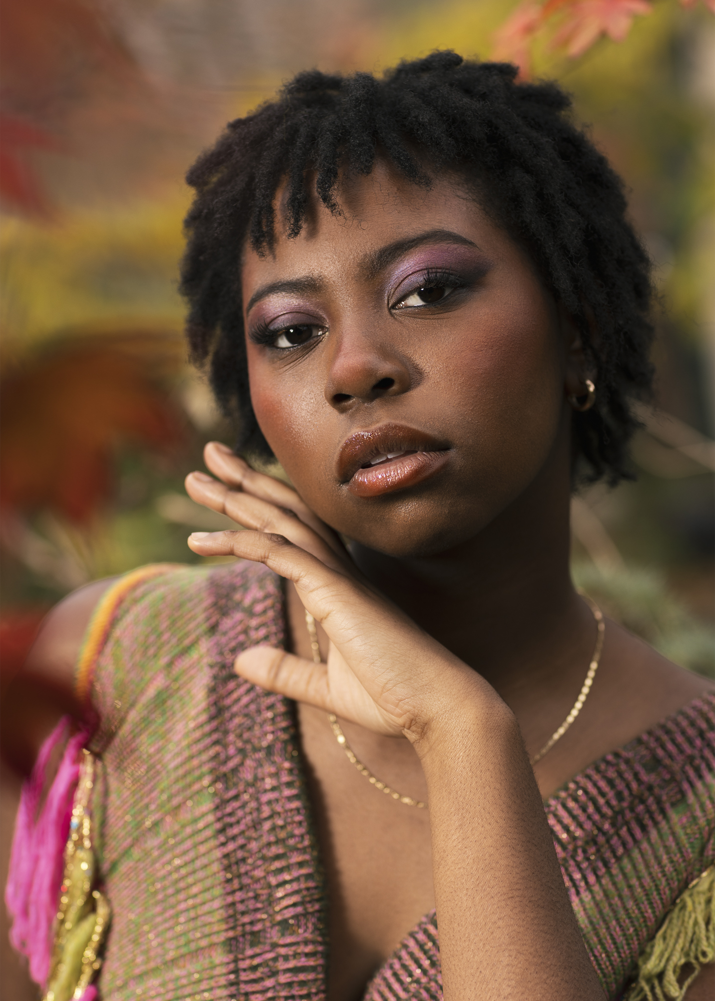A young woman with short, textured hair poses in front of autumn foliage. She wears a colourful, knitted top and has subtle makeup, with her left hand gently touching her chin as she looks towards the camera.