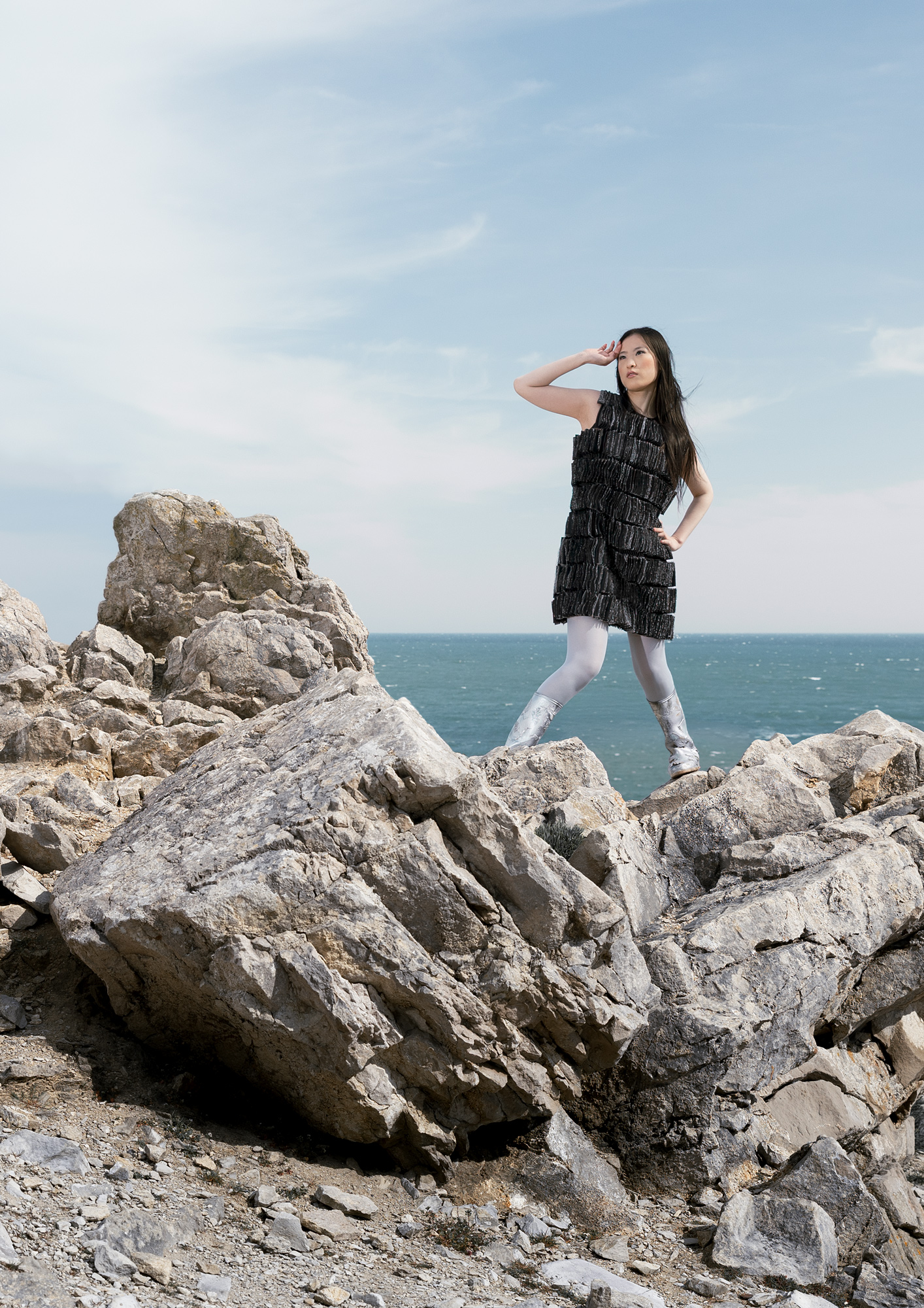 A woman wearing a black tiered dress stands on large rocks near the ocean. In the background, waves can be seen under a clear blue sky with scattered clouds.