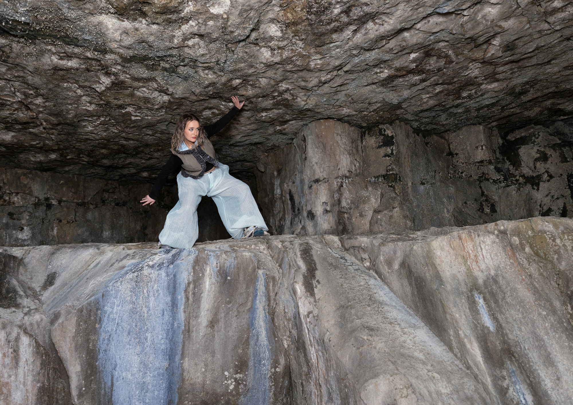 A person stands on a rocky ledge inside a cave, wearing a black top and light grey trousers. The cave features textured stone walls and a high ceiling, with natural light illuminating parts of the interior.