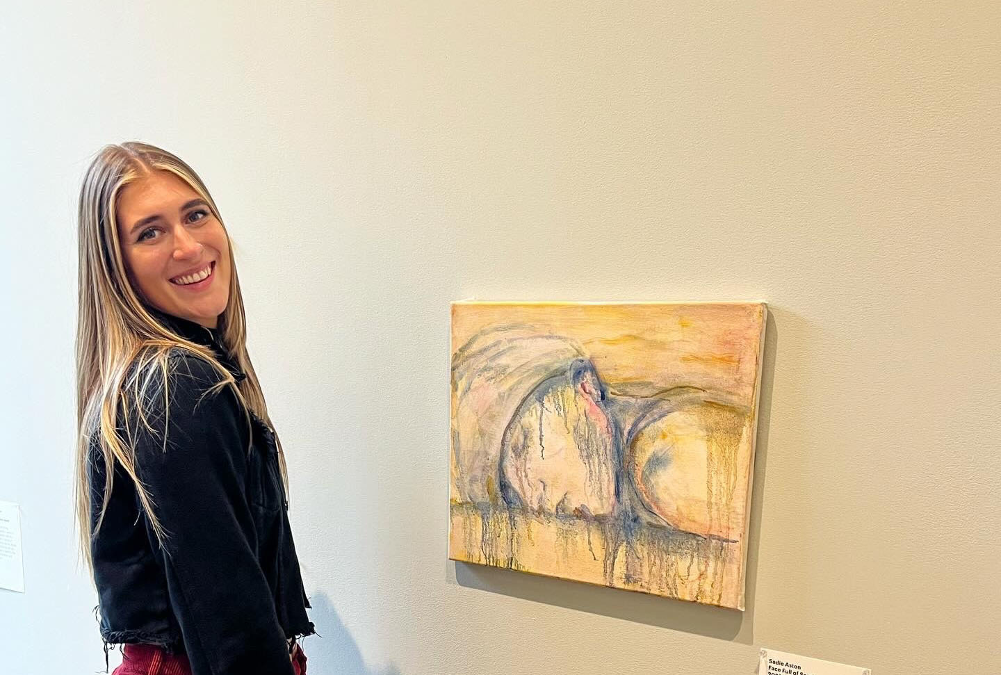 Blonde woman in black top stands beside framed artwork with yellow background and grey circular brushstrokes on white gallery wall.