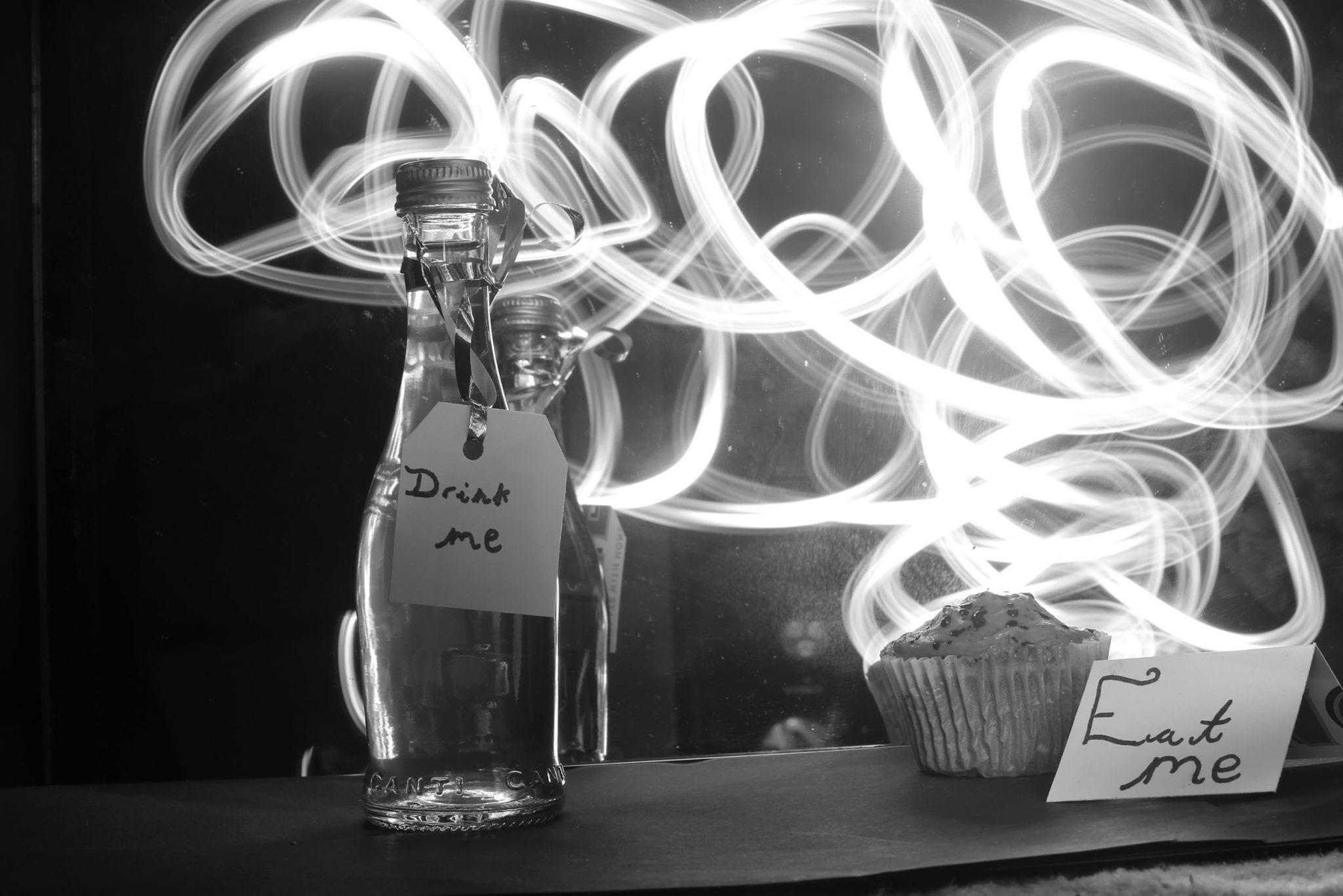 A glass bottle with a label reading "Drink me" and a cupcake with a label reading "Eat me". Bright light trails appear behind both objects.