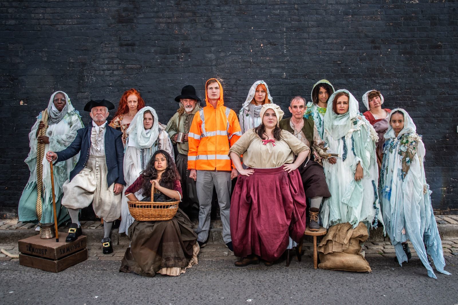 Cast photo of community play SALT, showing all performers in costume, posing for a photo in front of a black brick wall.