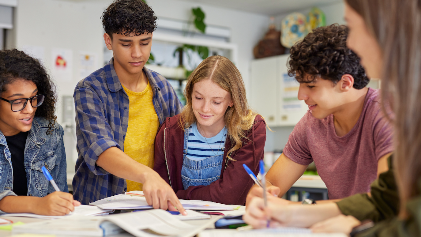 Five young people studying together at a table with papers and pens in a bright room with shelves