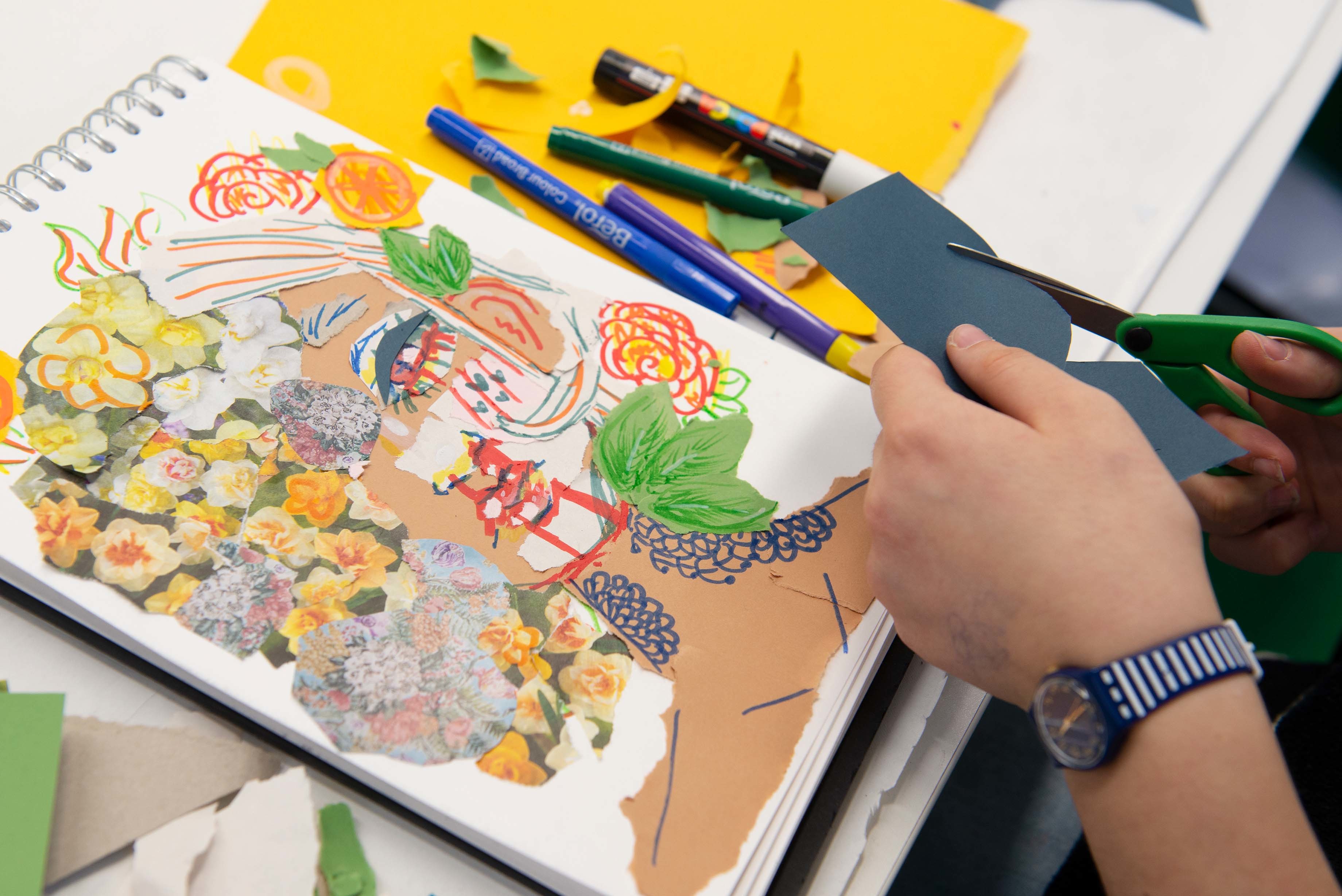 A person cuts shapes into a piece of card with a pair of scissors. A sketch pad sits next to them showing a colourful artwork of a human face half-covered by flowers.
