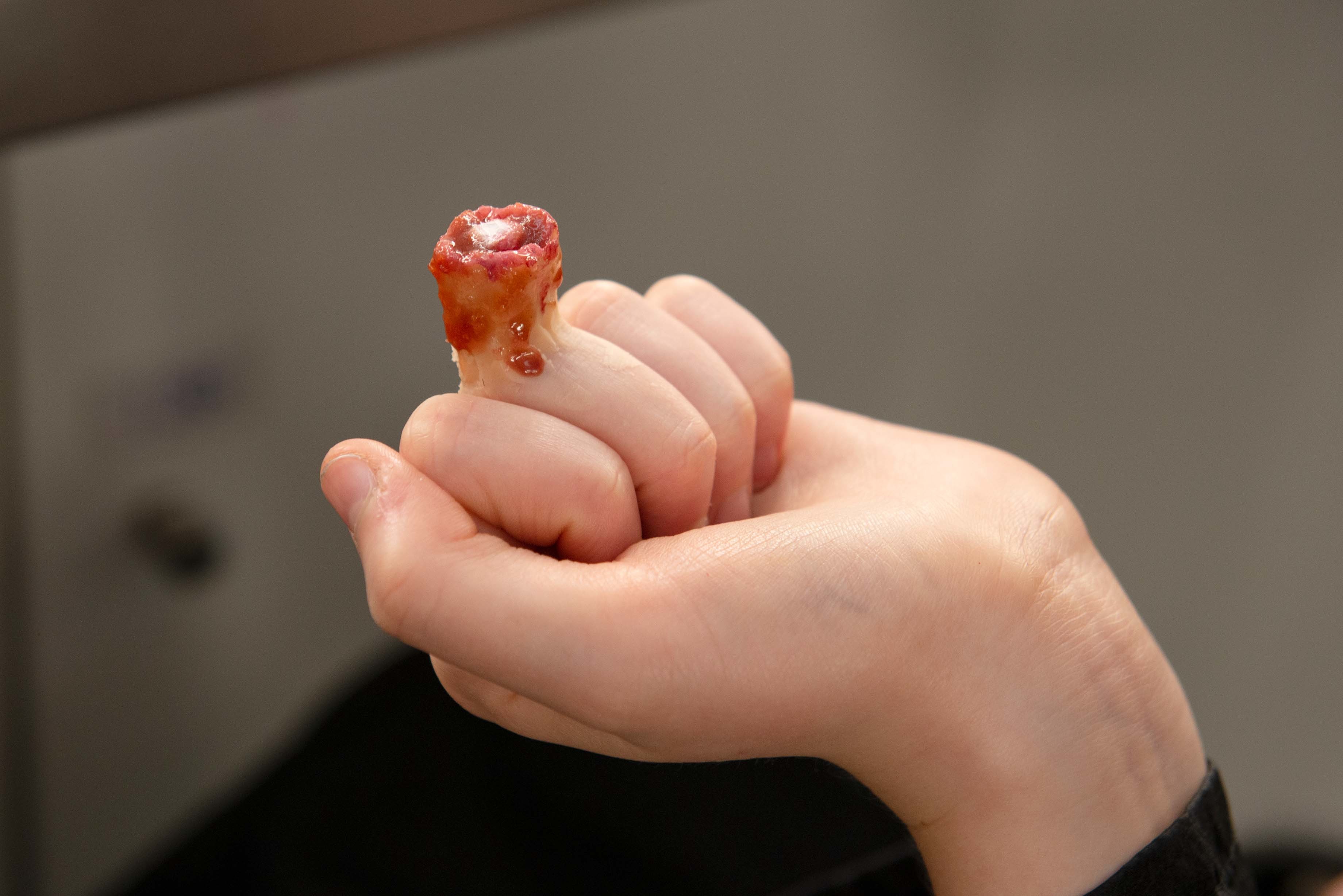 Hand holding small red translucent object between thumb and forefinger against blurred grey background.