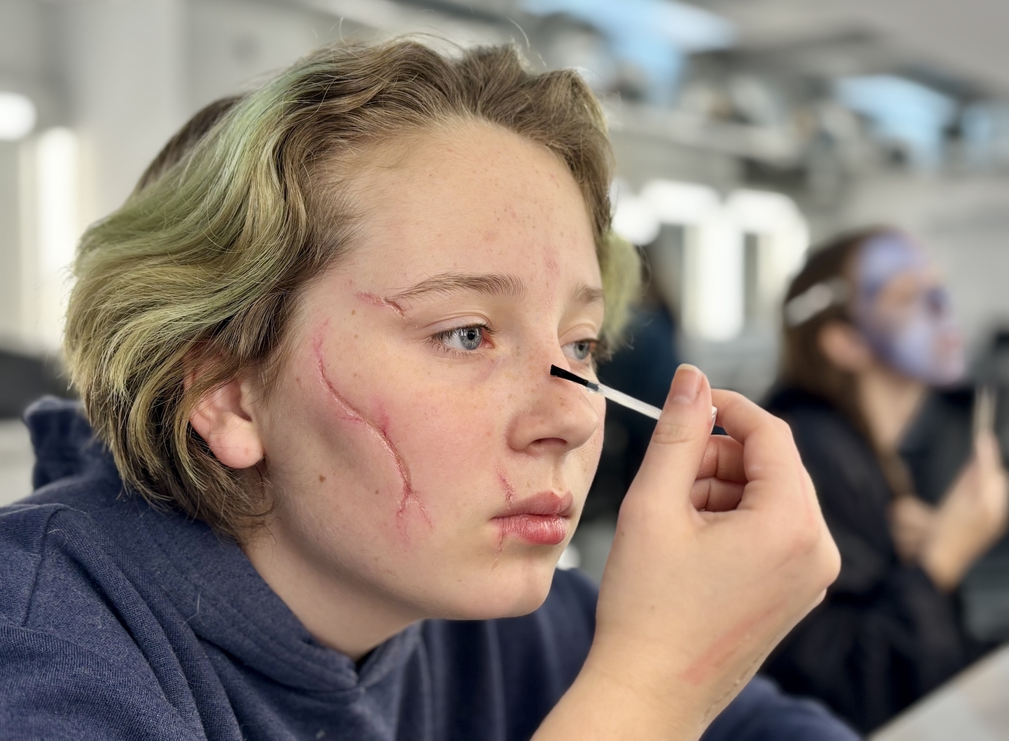Person with short blonde hair applying makeup with brush, red theatrical wounds painted on face, in studio setting