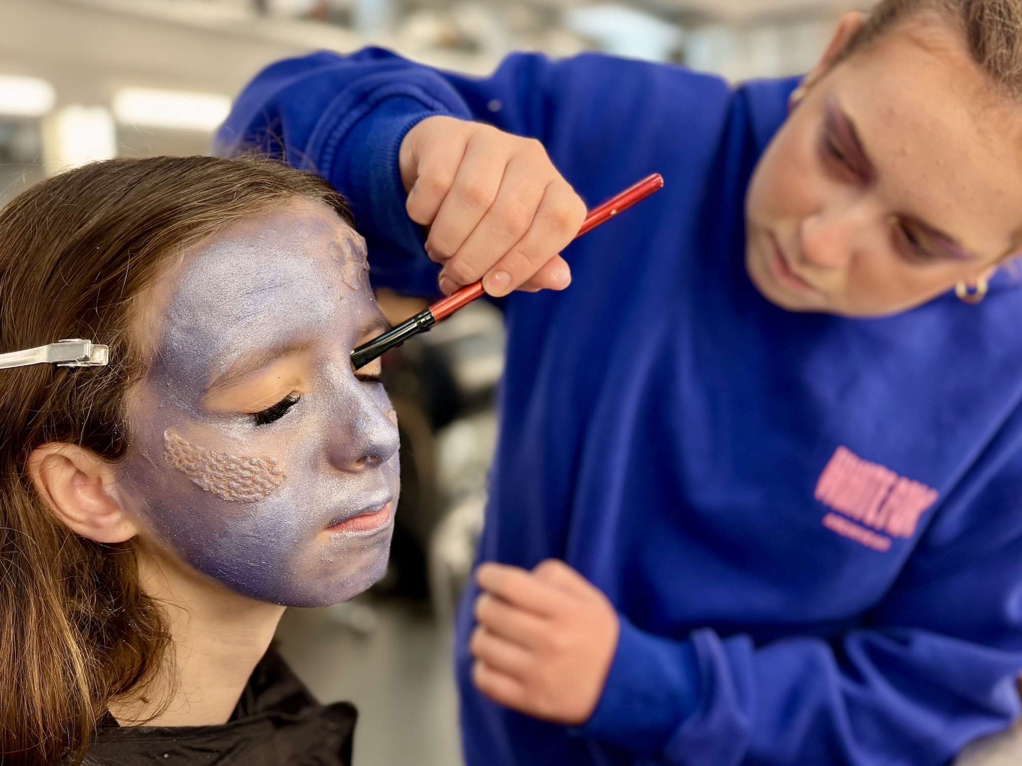 Make-up artist in blue jacket applying face paint to child with grey and orange designs on face using small brush