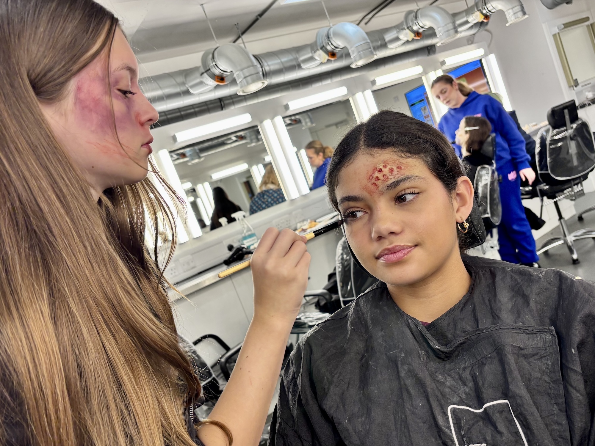 Makeup artist applying concealer to woman's face with brush in salon with mirrors and blue-uniformed staff in background