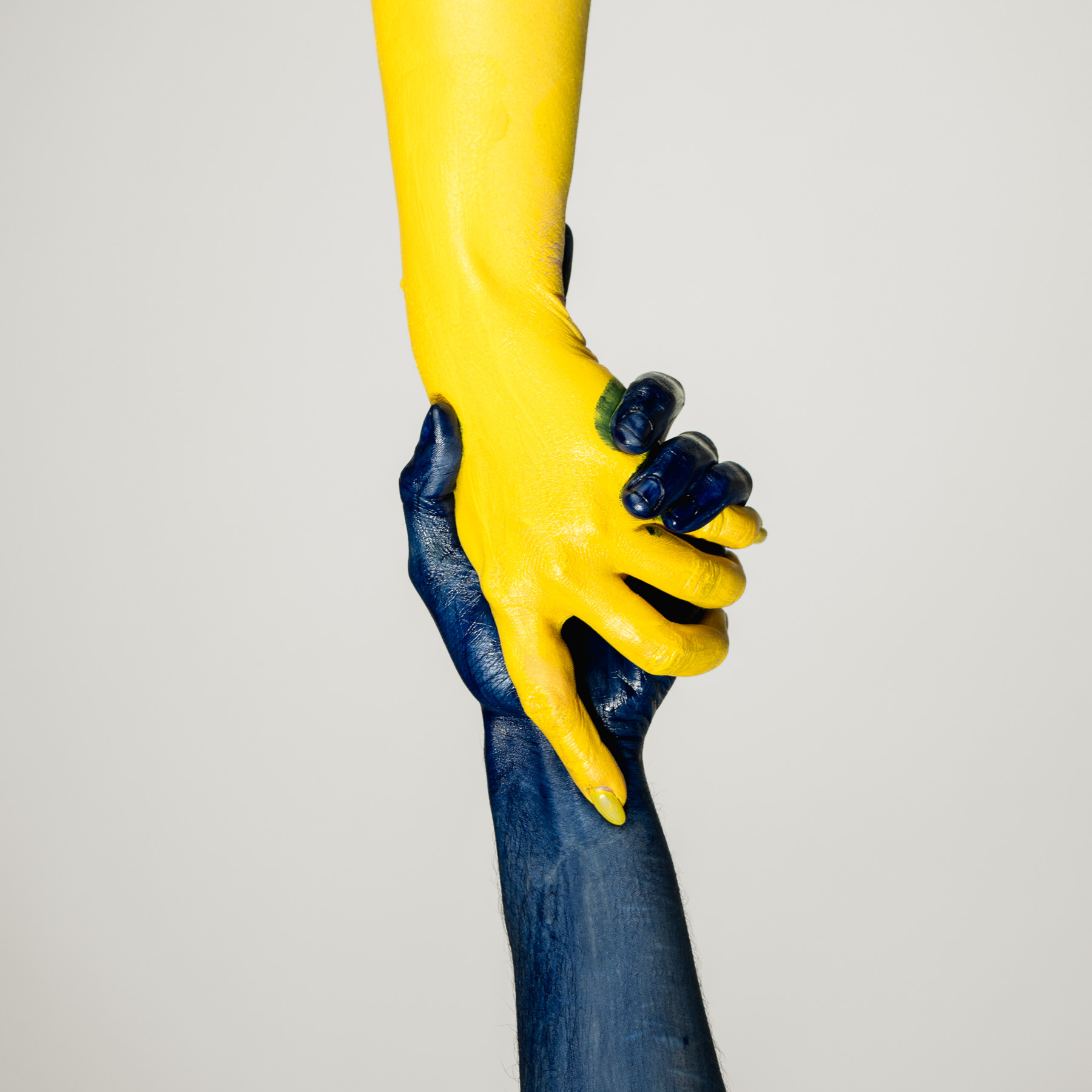 A close-up of two hands grasping each other, one hand is painted yellow and the other blue. The hands are positioned against a plain white background, and the grip is firm, with visible details of the paint on the skin.