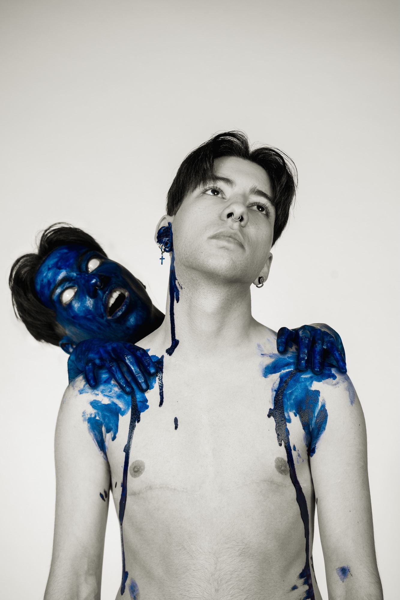 A man with short dark hair stands shirtless, adorned with blue paint on his skin. Another head, appearing to emerge from behind him, is also painted blue, with arms reaching around the man's shoulders. The background is neutral and plain.