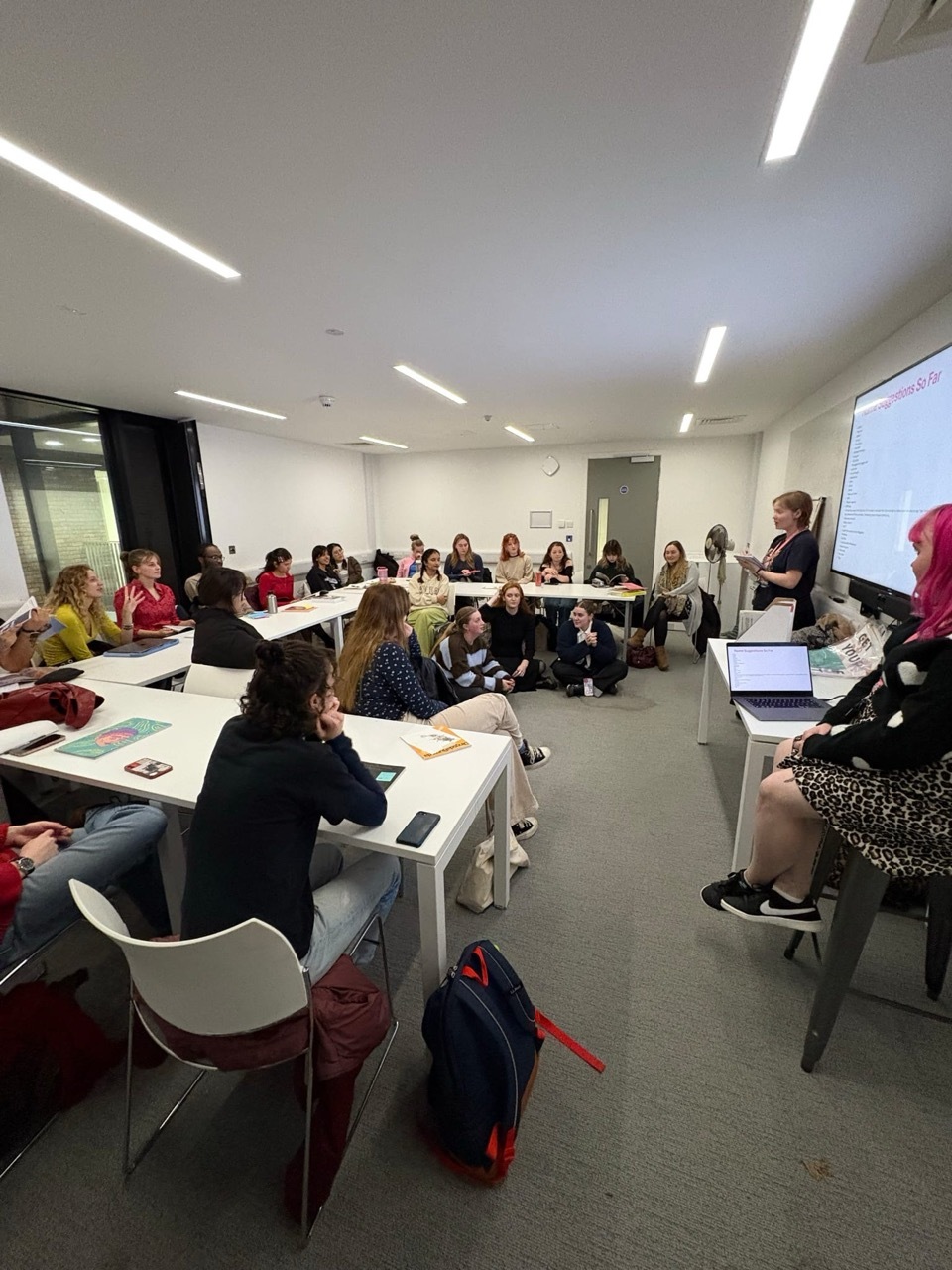 Modern classroom with white tables arranged in U-shape, students seated around perimeter, presenter at whiteboard, fluorescent lighting overhead.