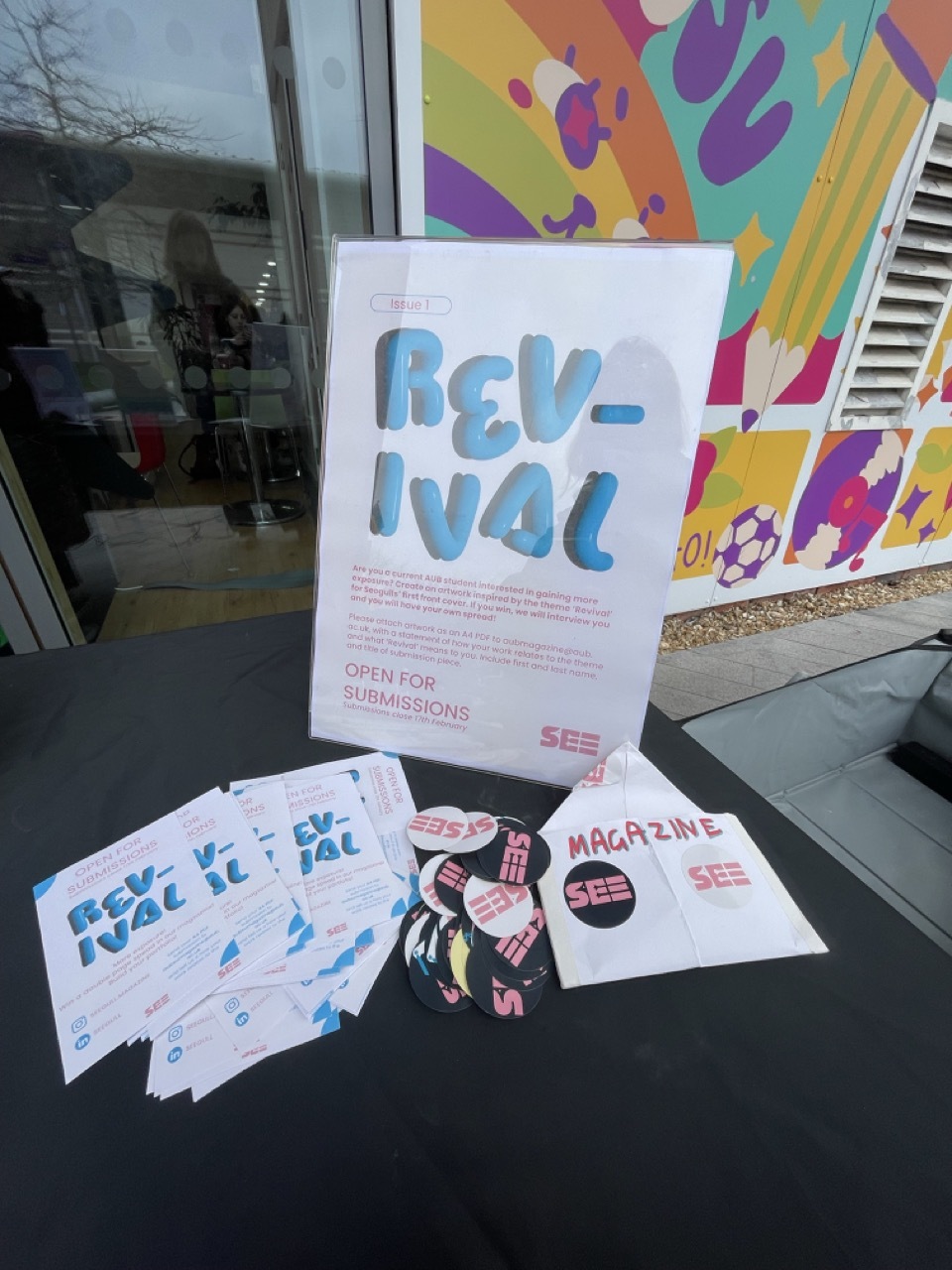 Revival poster with blue lettering on white paper displayed on glass surface, surrounded by printed materials and colourful wall mural.