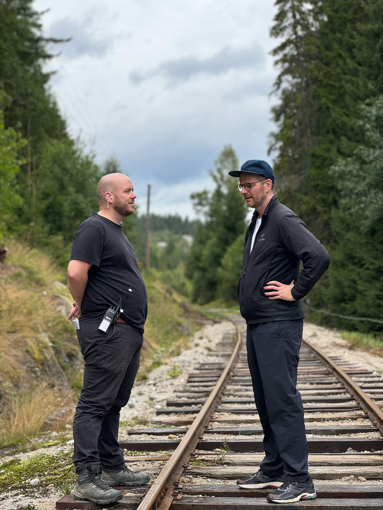 Two men in dark clothing standing on railway tracks having a conversation, surrounded by green coniferous trees under cloudy sky.
