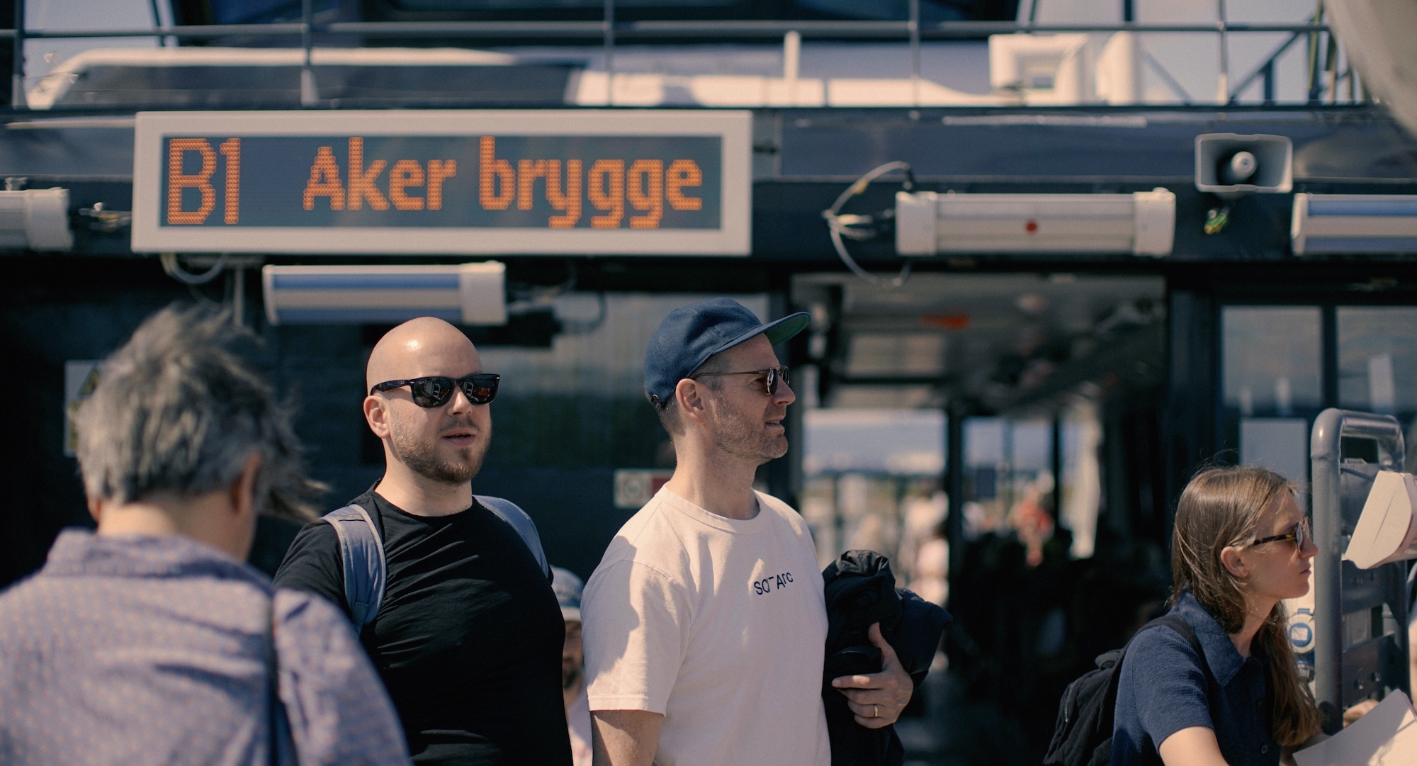 L T Skare and J Trier stand at Aker Brygge ferry terminal with Norwegian text sign above and passengers boarding boat in background.