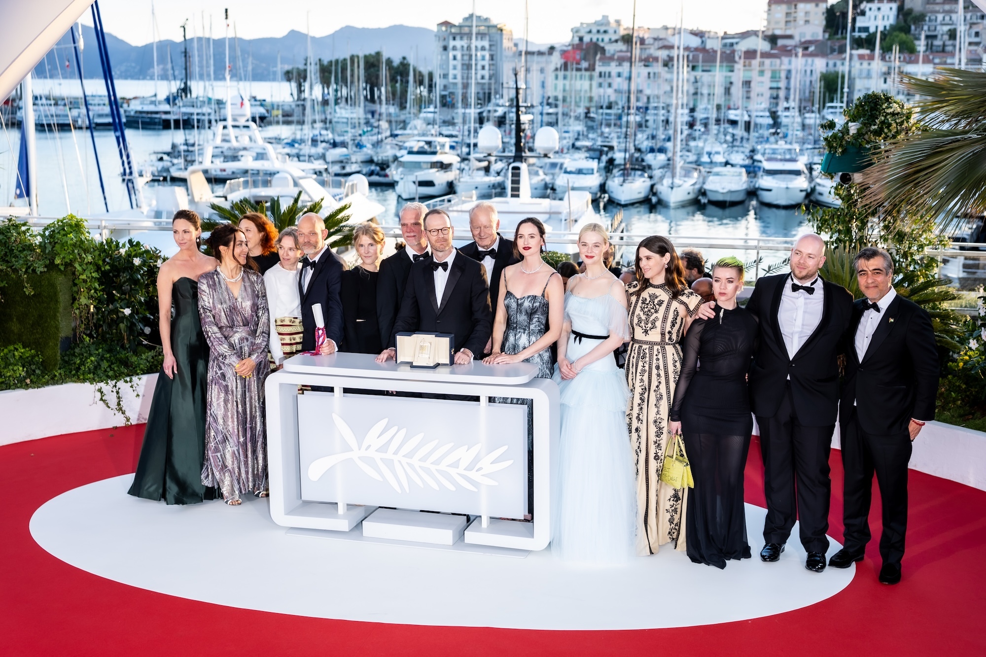 Cast and crew of "Sentimental Value" (2025) pose on red carpet at Cannes with award podium, marina and buildings visible in background.