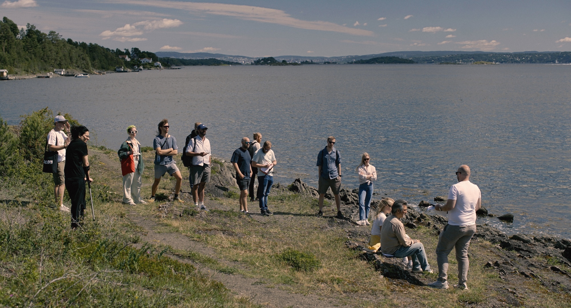 Group of people with tools working on rocky shoreline beside large lake, with forested hills and cloudy sky in background.